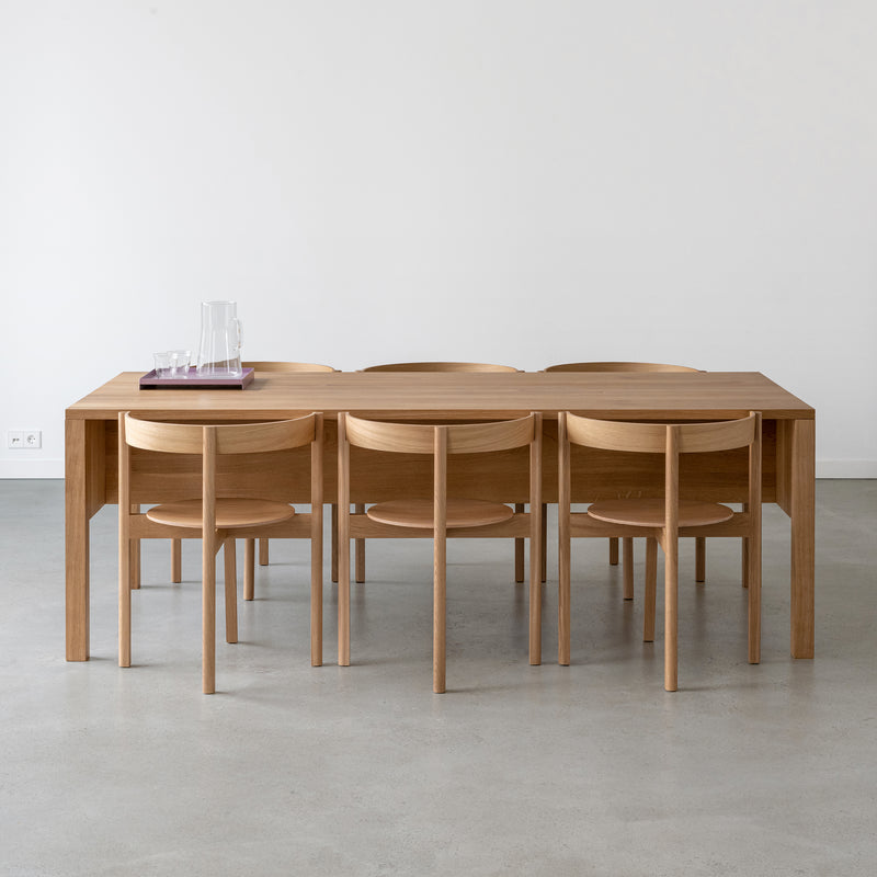 In a minimalist dining room, the L8 Traba Table by Loehr stands out, complemented by six matching chairs. A glass pitcher and four glasses on a purple tray grace the table. The serene ambiance is enhanced by diverse wood finishes, light gray floors, and white walls.