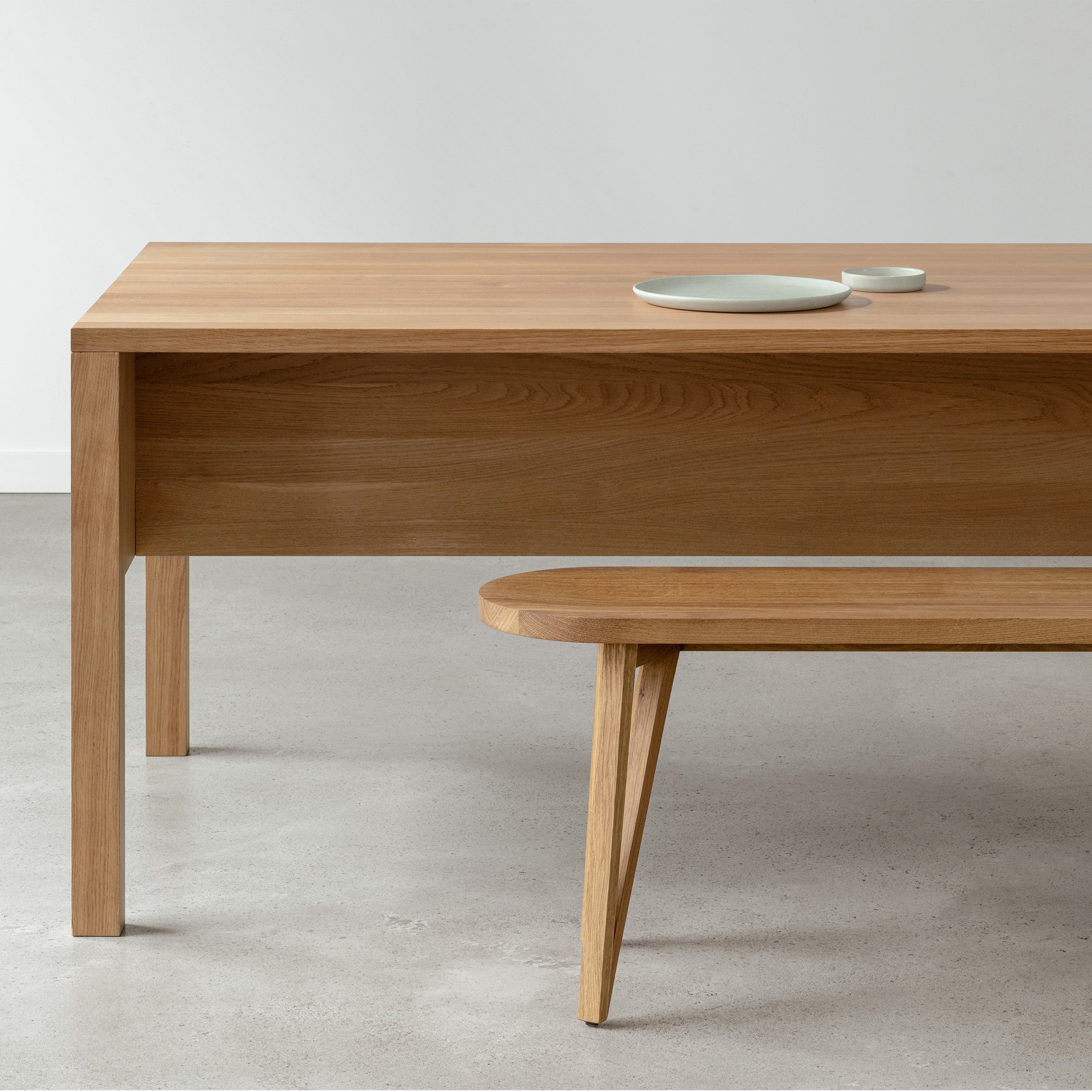 A minimalist dining area features the L8 Traba Table by Loehr and a matching bench on smooth concrete. Two small round ceramic plates are neatly placed, while the plain white wall in the background enhances the aesthetic appeal.