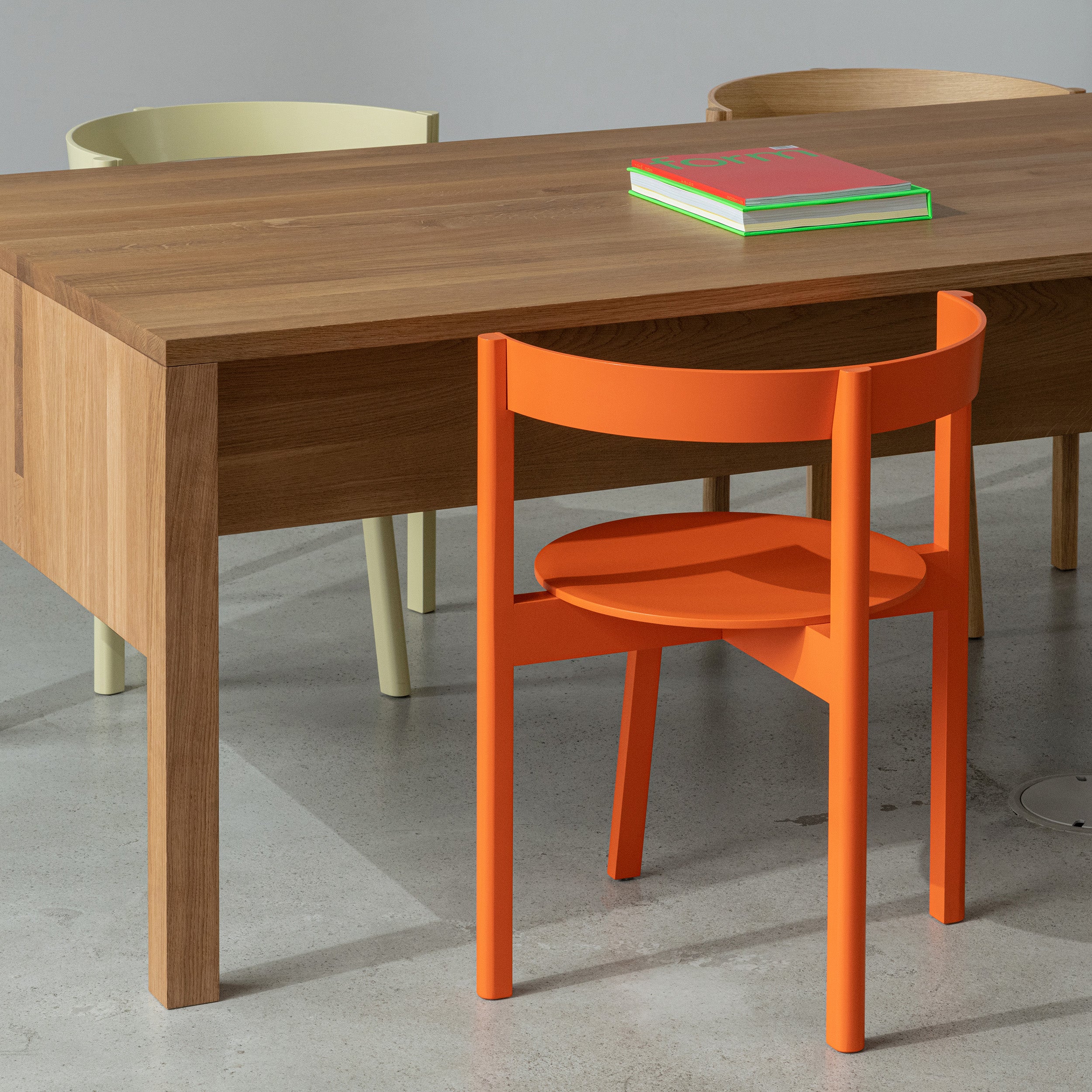 A minimalist room features the Loehr L8 Traba Table in solid wood, surrounded by two chairs in mixed wood finishes and one bright orange chair. A green book sits atop the table, while the gray concrete floor complements this modern aesthetic.