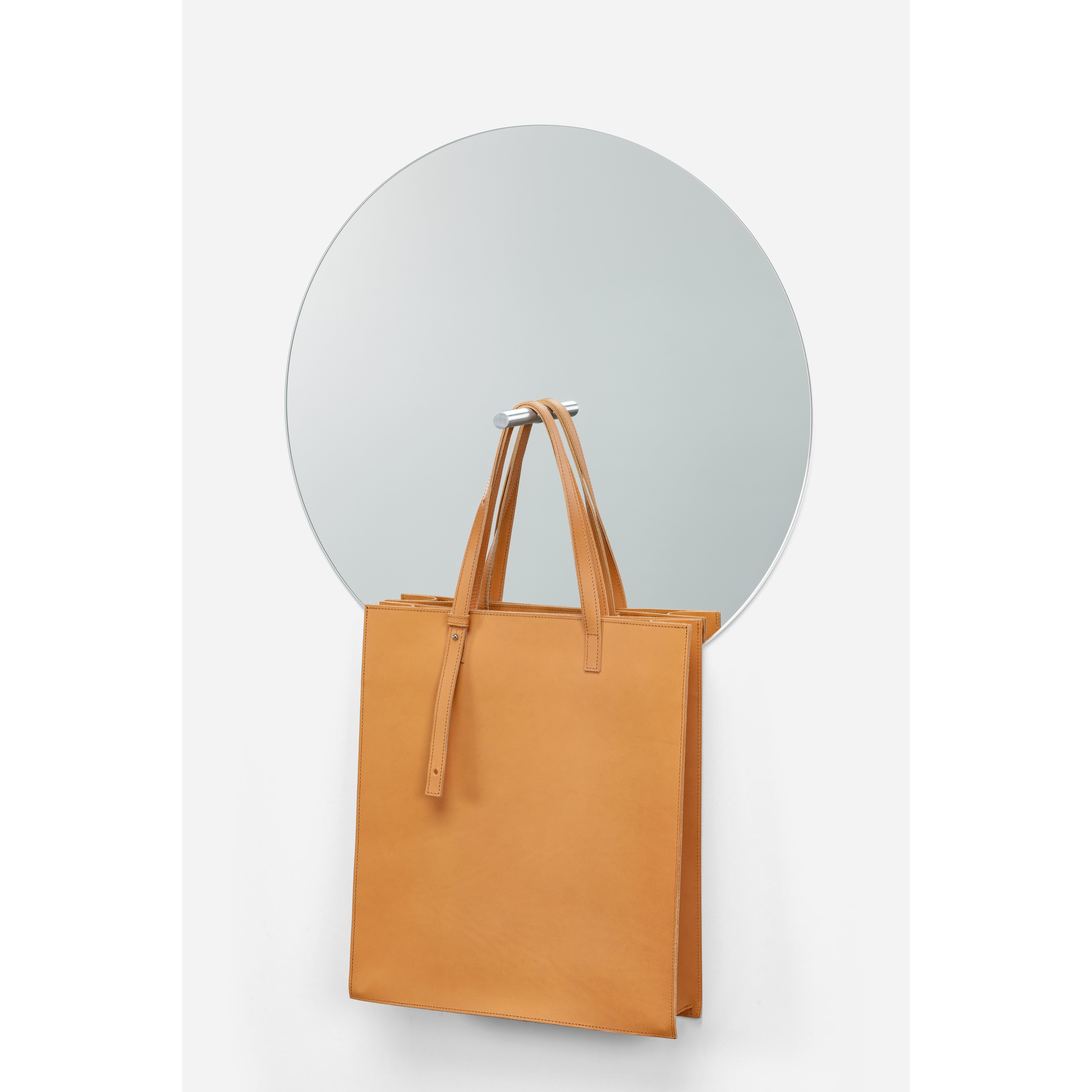 A brown leather tote bag with two handles hangs on a round, frameless L9 Portal mirror by Loehr against a plain white background, transforming the mirrored surface into an elegant decorative piece.