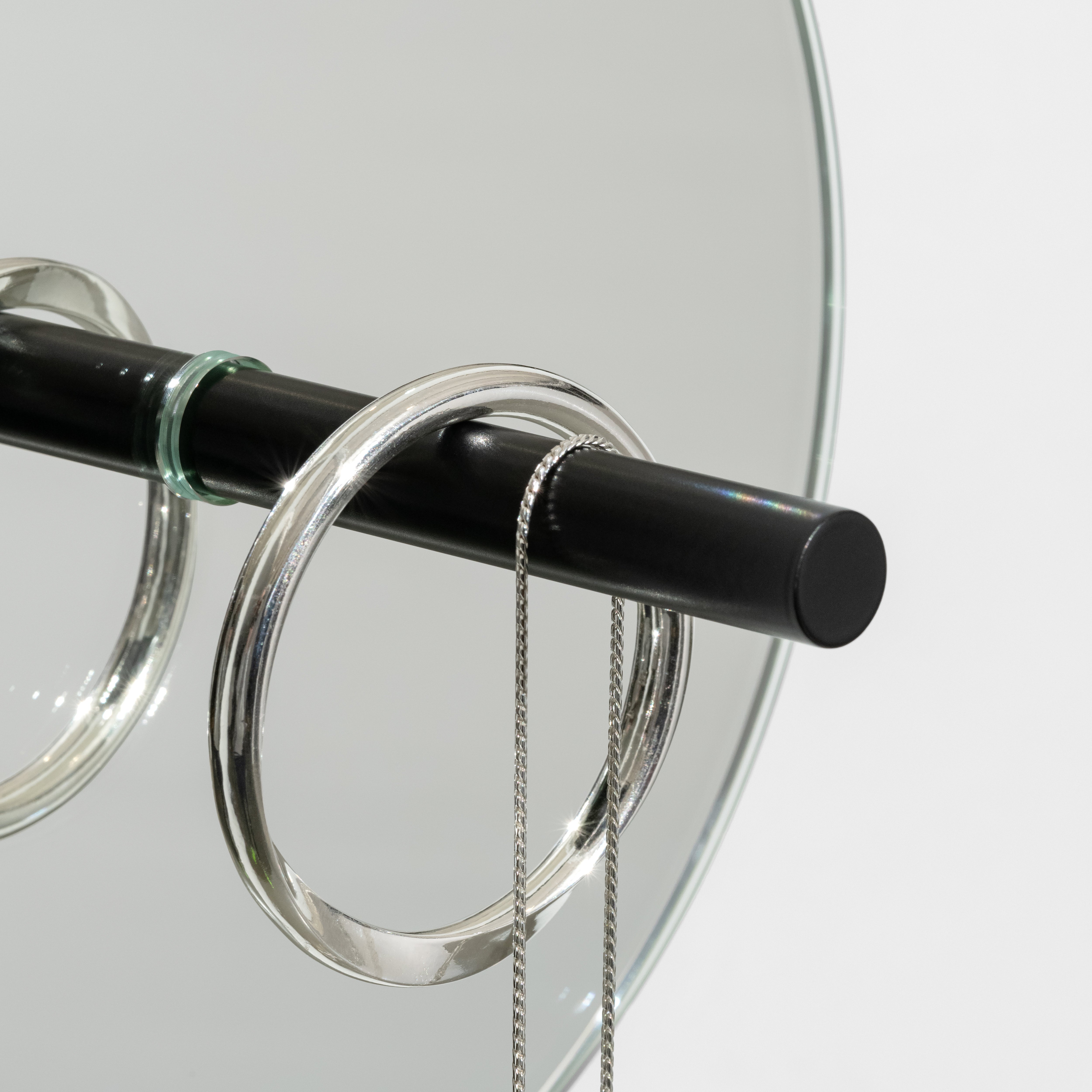 The L9 Portal by Loehr features a black rod with circular metal rings, resembling a minimalist jewelry stand. A thin silver chain drapes elegantly from the rod, while the mirrored background surface adds depth to this decorative and functional piece.