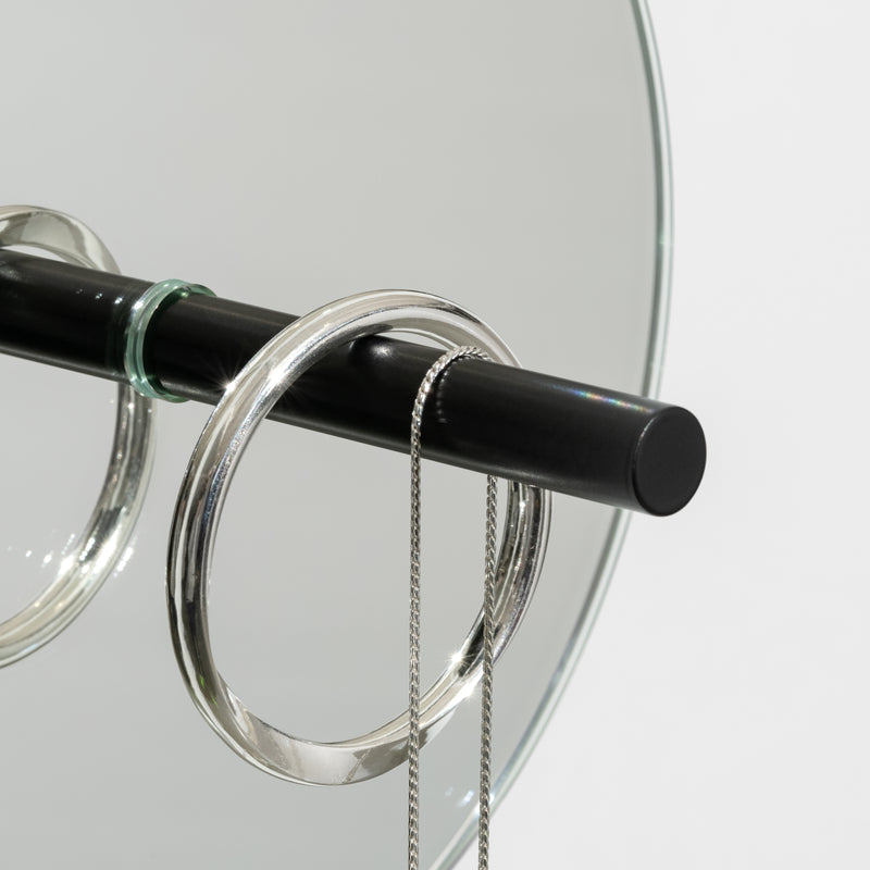 The L9 Portal by Loehr features a black rod with circular metal rings, resembling a minimalist jewelry stand. A thin silver chain drapes elegantly from the rod, while the mirrored background surface adds depth to this decorative and functional piece.