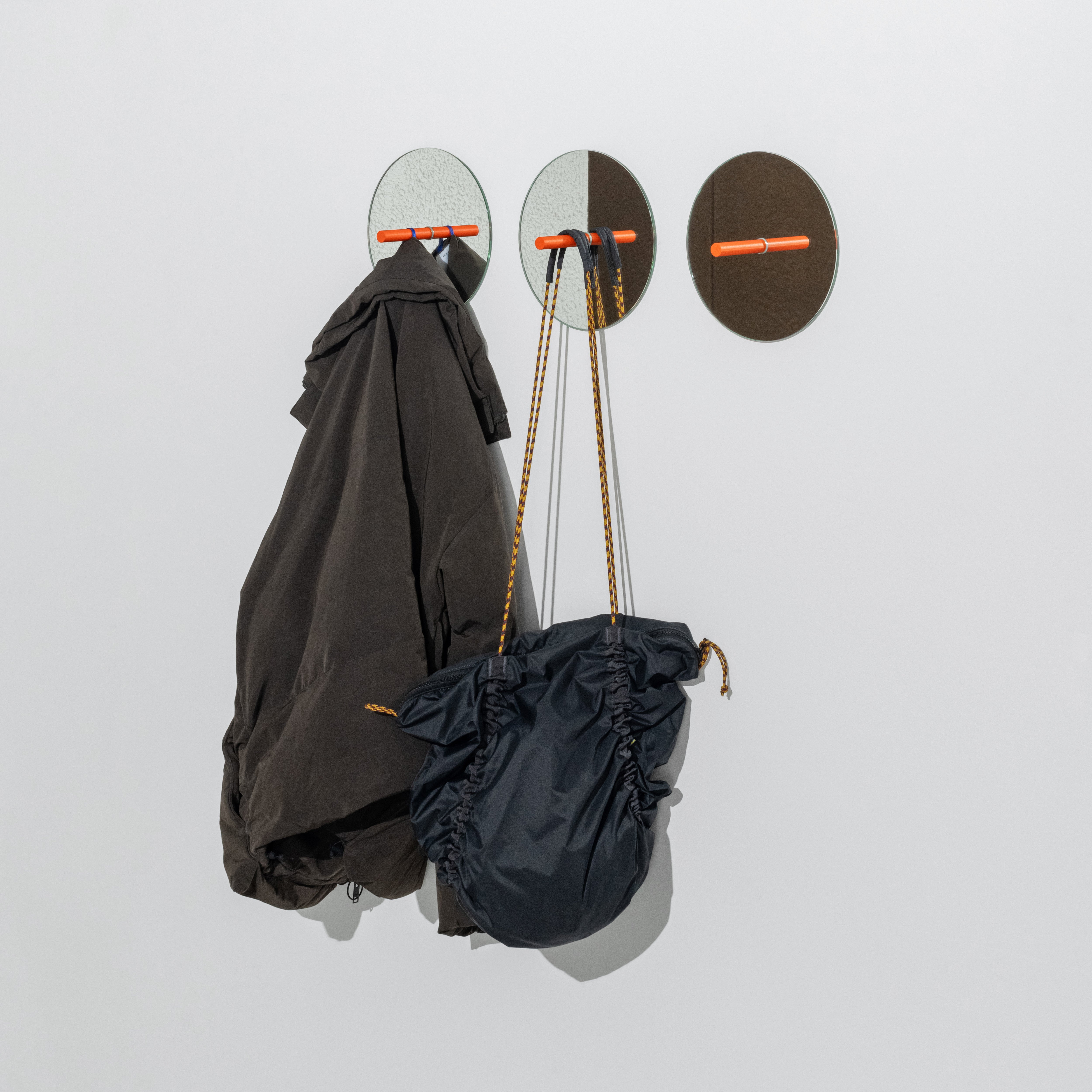 The L9 Portal by Loehr features three round wall hooks with mirrored surfaces and orange pegs, showcasing a minimalist design with a dark jacket and bag hanging against a plain white background to create a modern look.