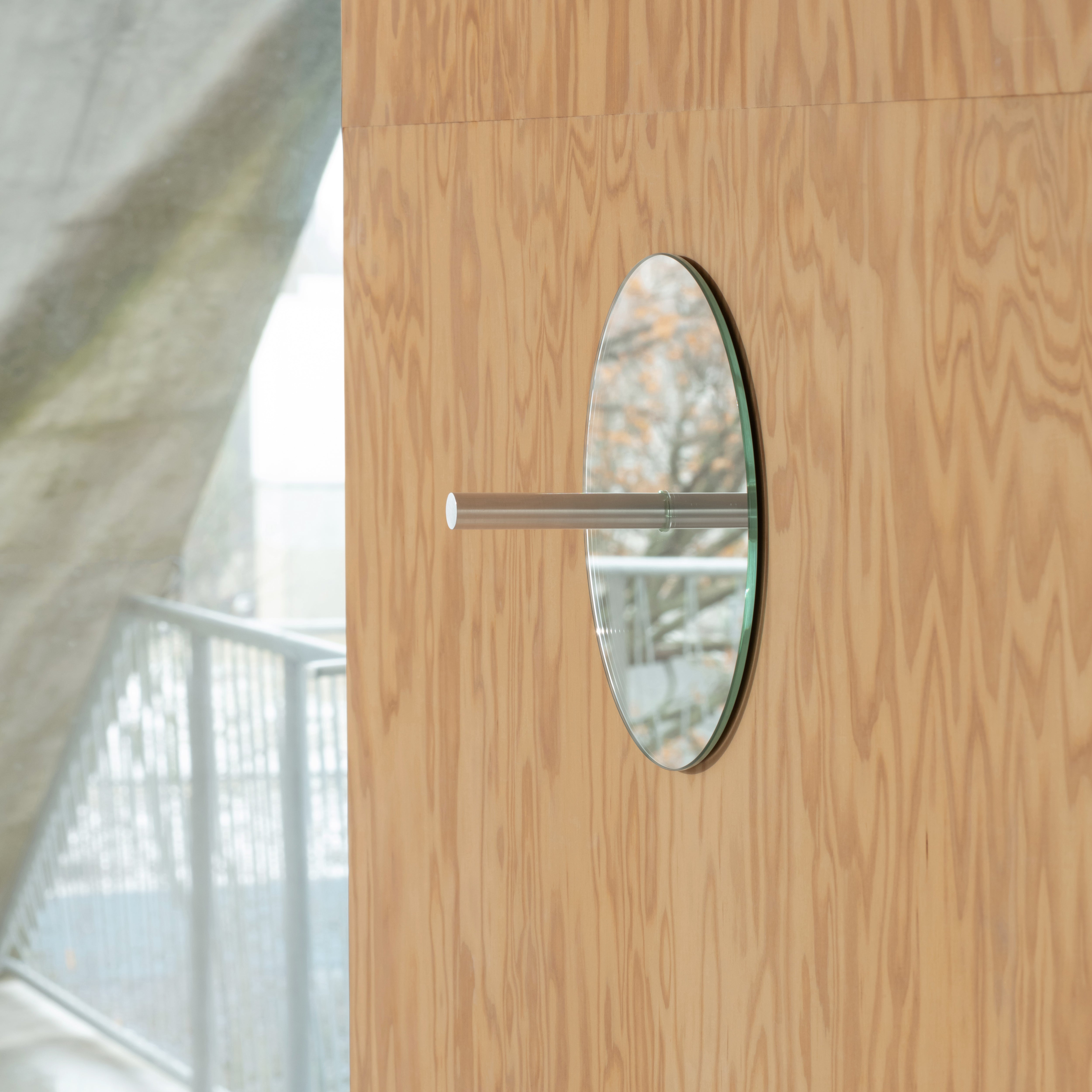 The L9 Portal by Loehr is a minimalistic, wall-mounted circular mirror with a cylindrical detail. It reflects an outdoor scene featuring a metal rail and blurred nature against plywood, serving both as decoration and function.
