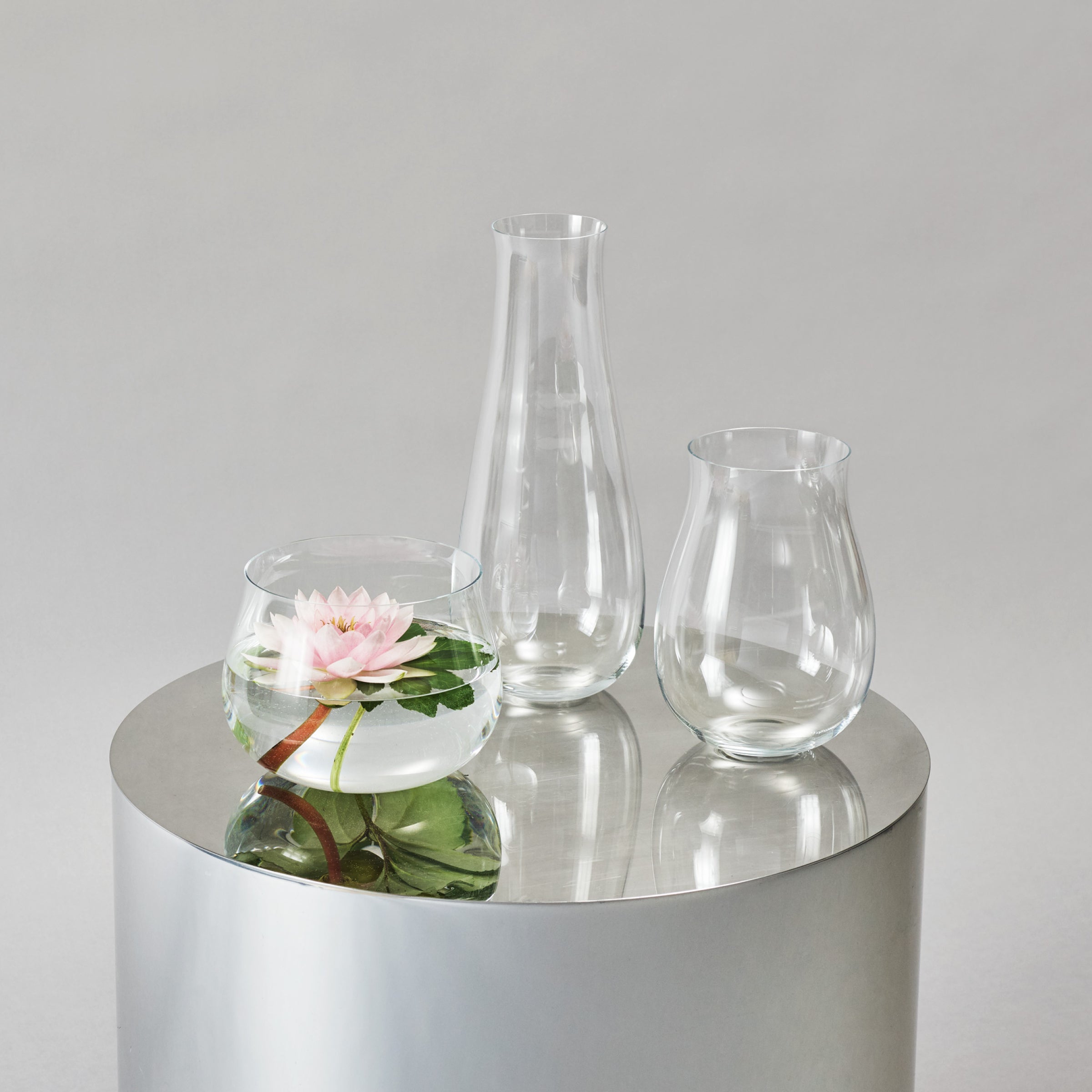 Three Lobmeyr BV69 Drop Vases sit on a reflective circular surface; the left vase holds water and a pink lotus flower, showcasing exquisite flower arranging, while the center vase is tall and narrow, with the right one being slightly shorter and wider. The background is light gray.