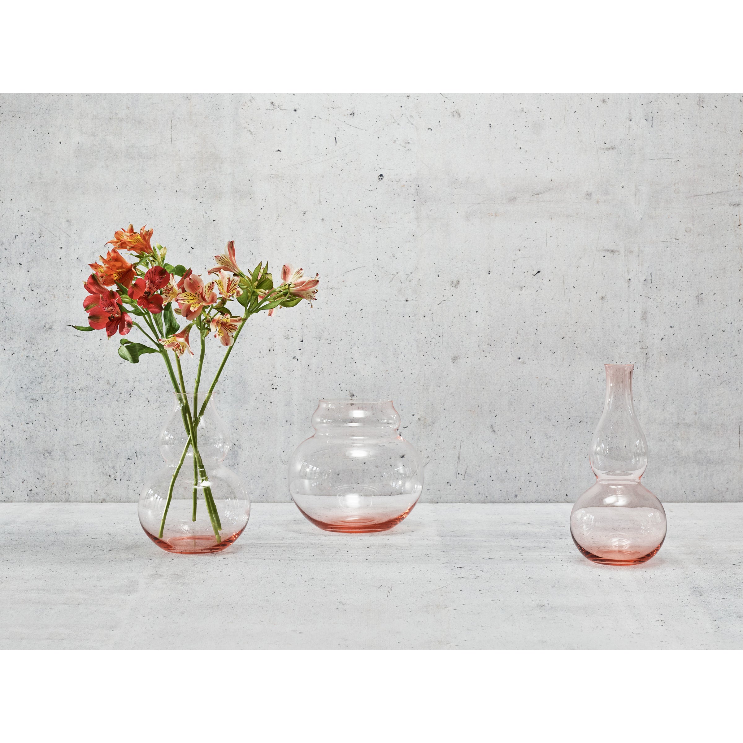 On a light gray concrete surface, three mouth-blown, lead-free crystal vases from Lobmeyrs BV71 Calabash Flower Vase line showcase their elegance. The left contains orange flowers, the middle is round and open, while the right is narrow with a wavy design. All have a subtle bottom orange tint.