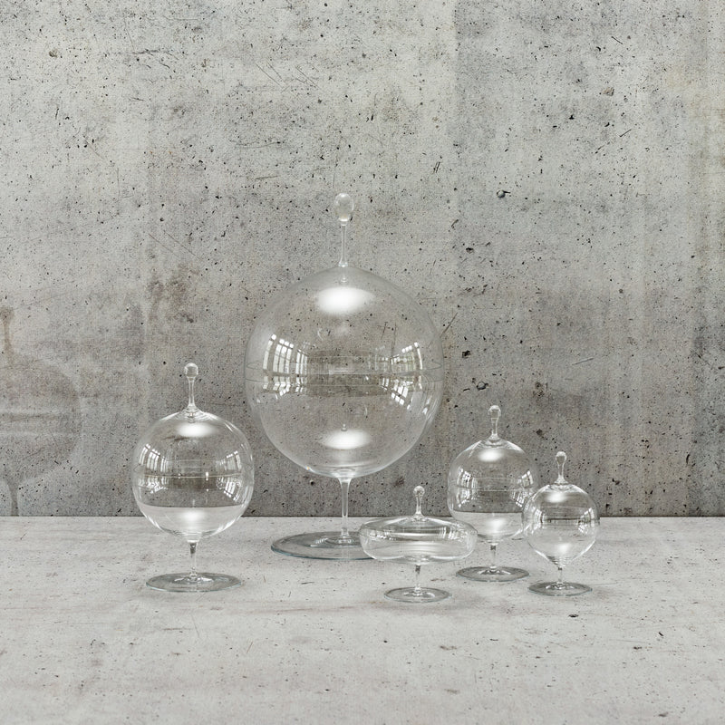 A set of five mouth-blown crystal containers inspired by Oswald Haerdtls design stands against a concrete backdrop. These spherical Candy Dishes by Lobmeyr reflect light brilliantly, featuring elegant pedestal bases reminiscent of classic styles.