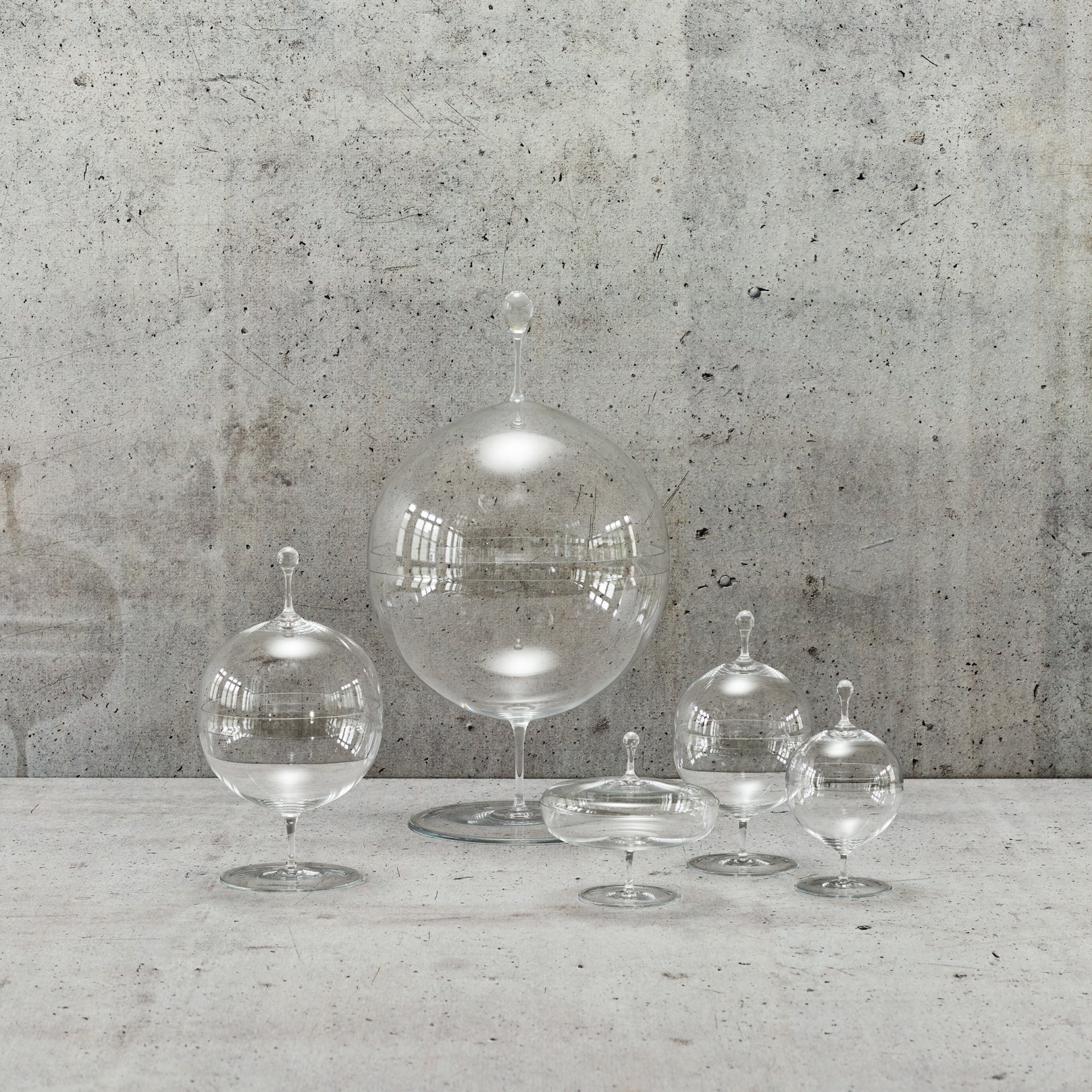A set of five clear glass cloches, reminiscent of Oswald Haerdtls style and crafted from mouth-blown lead-free crystal by Lobmeyr, are elegantly placed on a textured concrete surface. Their pointed tops beautifully reflect light, creating bright highlights against the rough gray concrete wall.