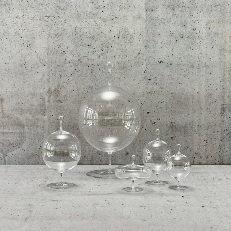 A set of five clear glass cloches, reminiscent of Oswald Haerdtls style and crafted from mouth-blown lead-free crystal by Lobmeyr, are elegantly placed on a textured concrete surface. Their pointed tops beautifully reflect light, creating bright highlights against the rough gray concrete wall.