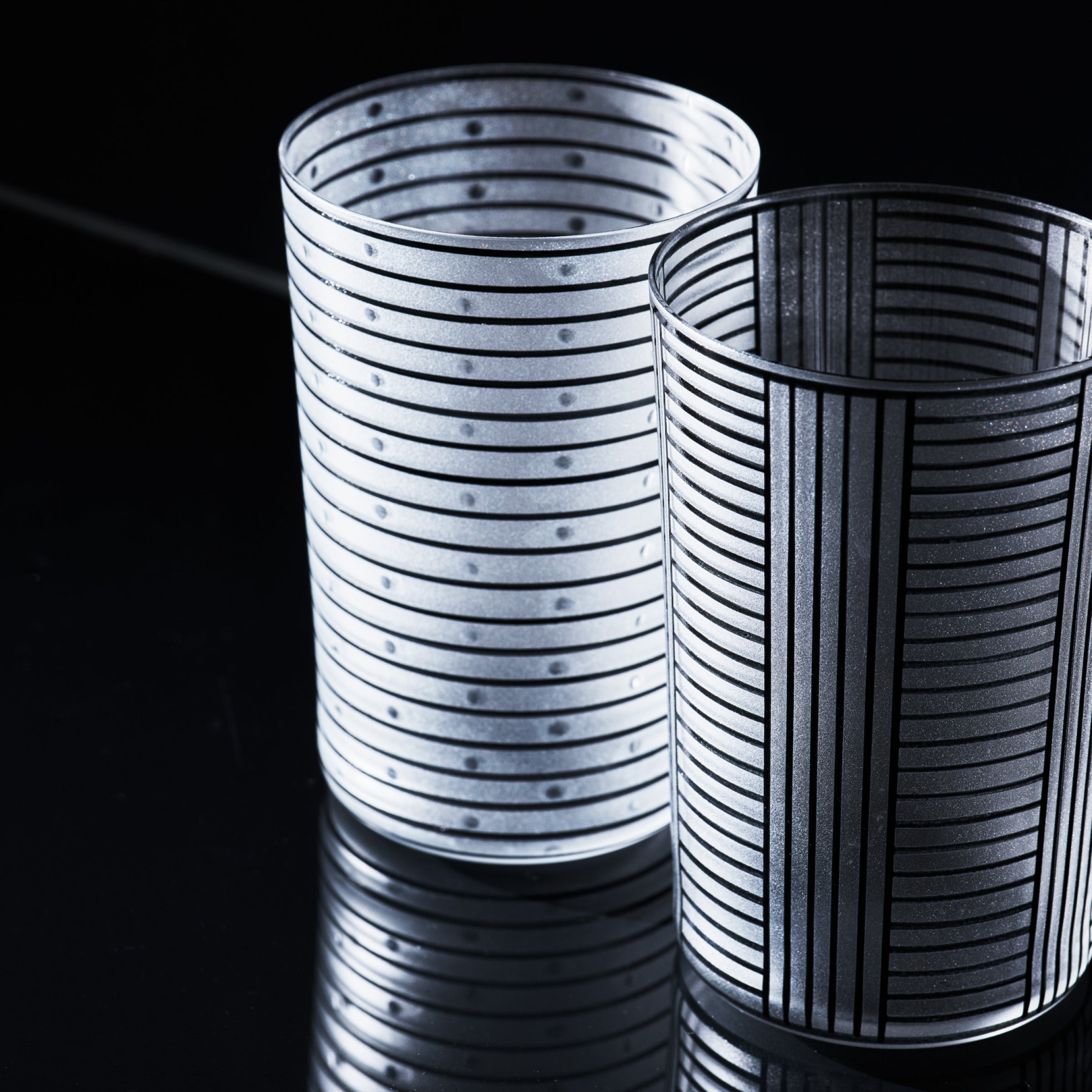 Two lead-free crystal glasses from the Lobmeyr Drinking Set Serie A, featuring vertical and horizontal black stripes, sit side by side on a reflective black surface, their patterns casting light and shadow to enhance contrast against the dark background.