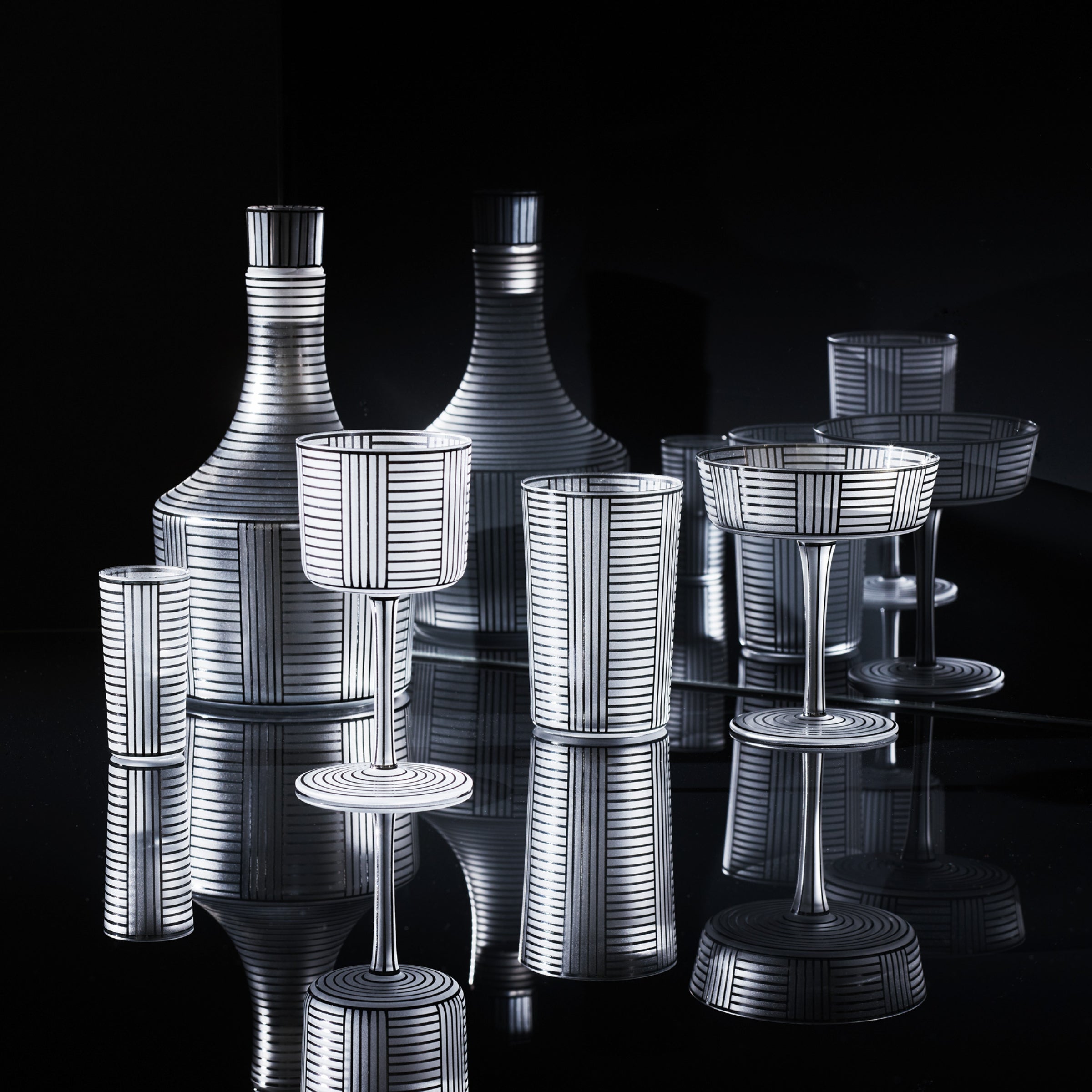 The Drinking Set Serie B by Lobmeyr features elegant crystal glassware with geometric white line patterns. The collection includes mouth-blown decanters, goblets, and tumblers. Displayed against a black background, they evoke the intricate reflections of Josef Hoffmann’s design style.