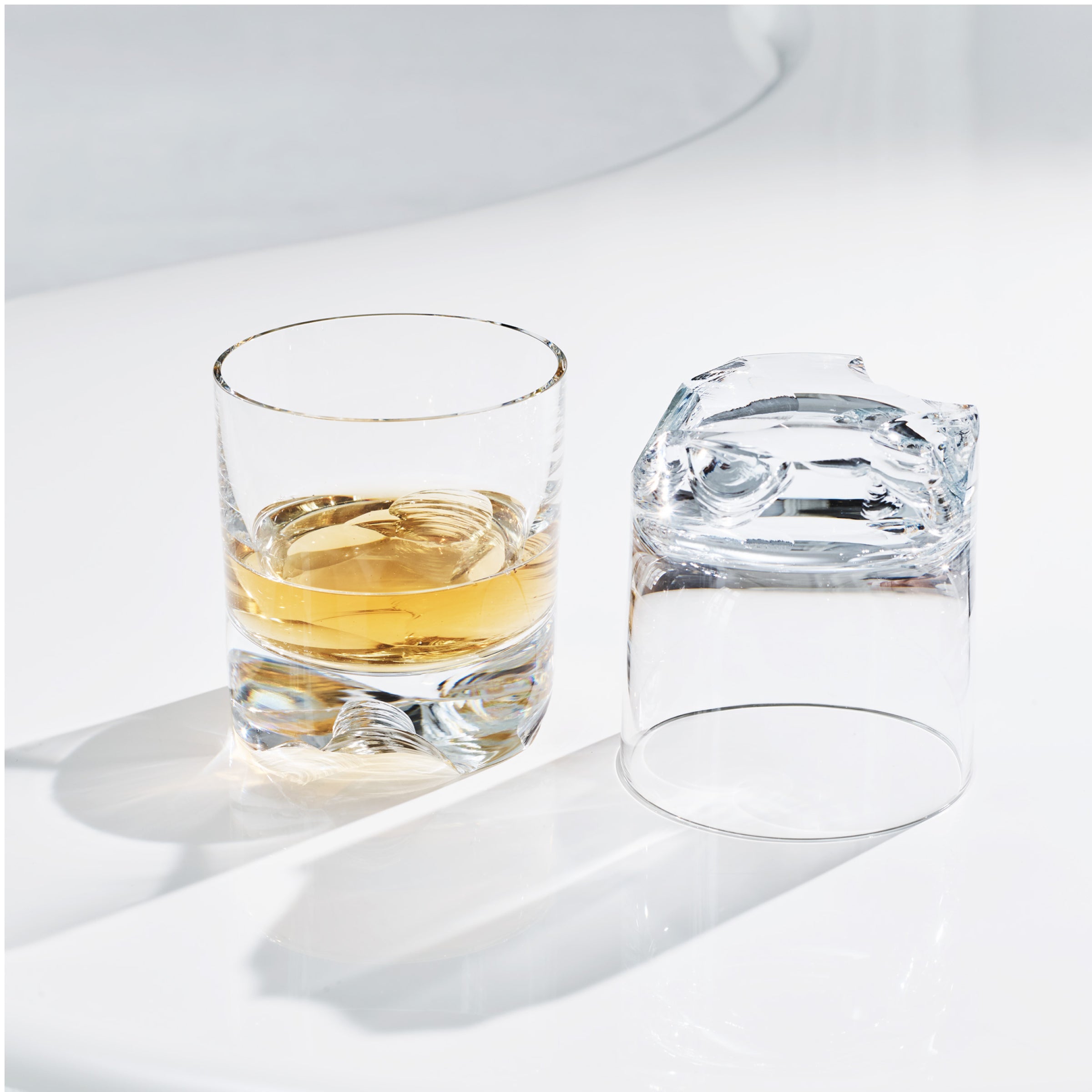 A clear Lobmeyr Bar Series - Flint glass filled with golden liquid sits on a smooth white surface, while another identical glass lays on its side, casting a distinct shadow. Bright lighting emphasizes the minimalistic scene and highlights the glass and liquid.