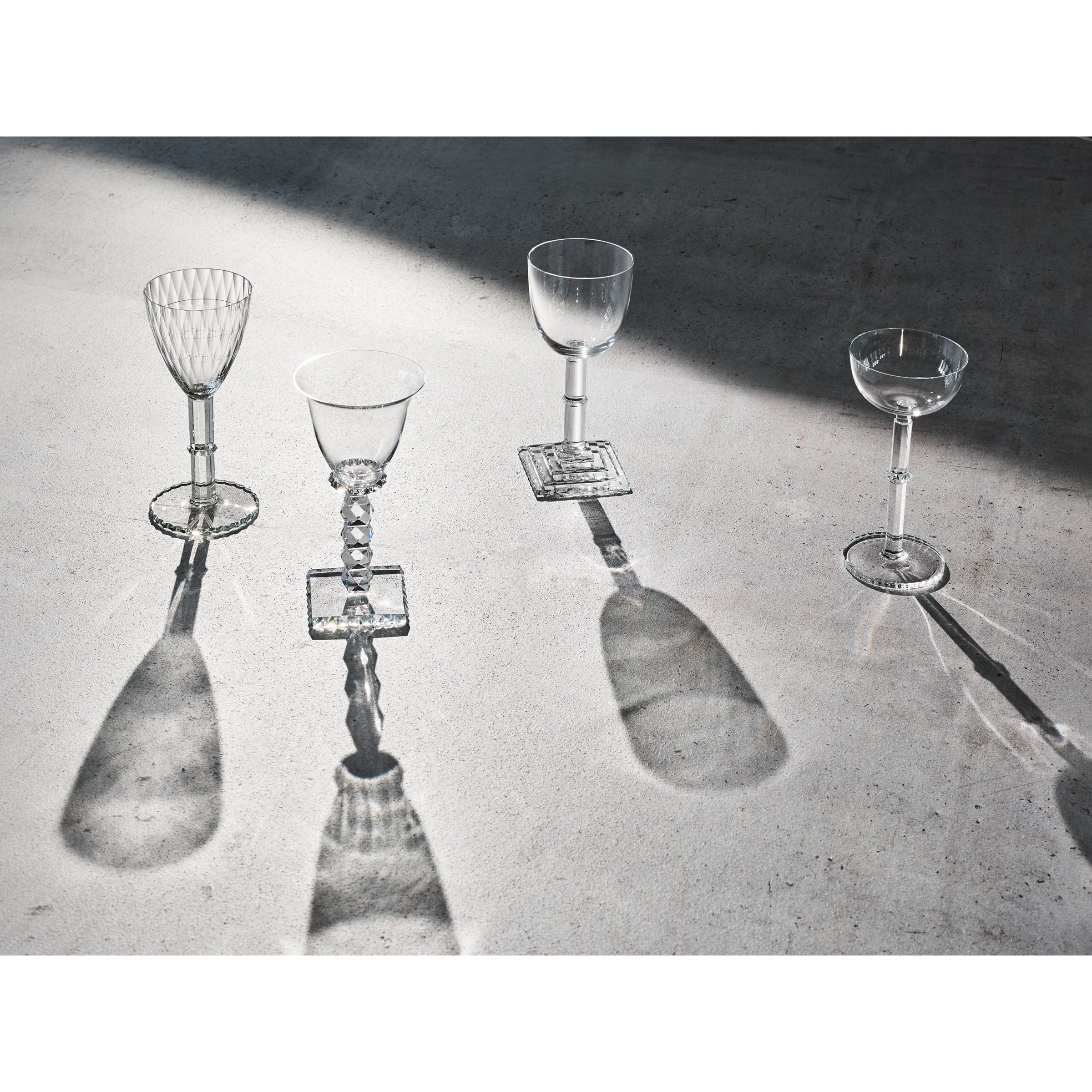 Four empty Hoffmann Goblets by Lobmeyr, crafted from mouth-blown lead-free crystal, feature unique stem and bowl designs reminiscent of Josef Hoffmanns style. They cast artistic shadows on a textured, light-colored surface under soft, natural light.