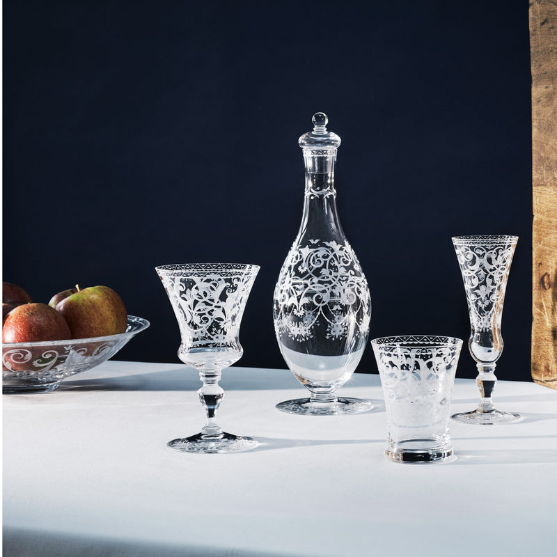 The Lobmeyr Drinking Set No. 231 - Barock - with engraved ornament, displayed on a white cloth against a dark backdrop, features an ornate carafe, two hand-engraved wine glasses, and a tumbler. A bowl of apples is partially visible on the left, with dramatic top lighting evoking Viennese Baroque artistry.