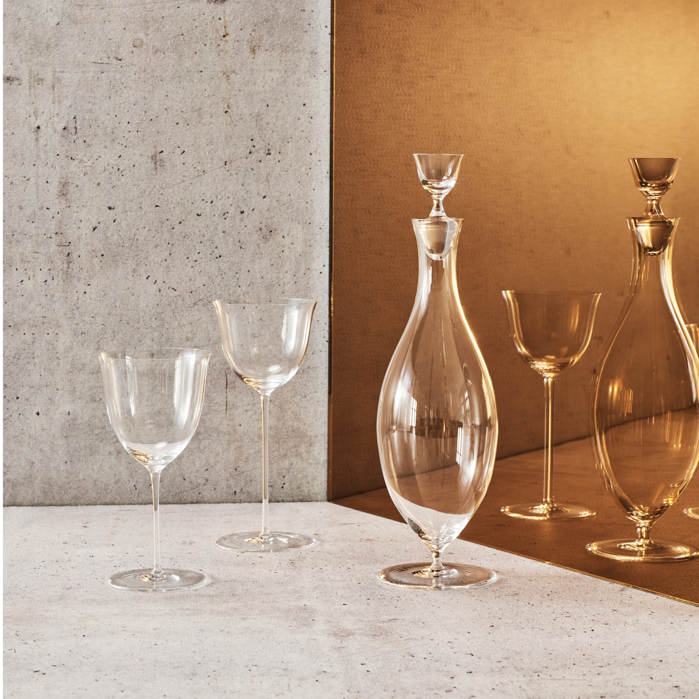 The Drinking Set No. 238 - Patrician by Lobmeyr, featuring a glass decanter and two wine glasses made from mouth-blown crystal, sits on a concrete surface beside a mirrored wall. The decanter and glasses create an elegant symmetry with their reflection.