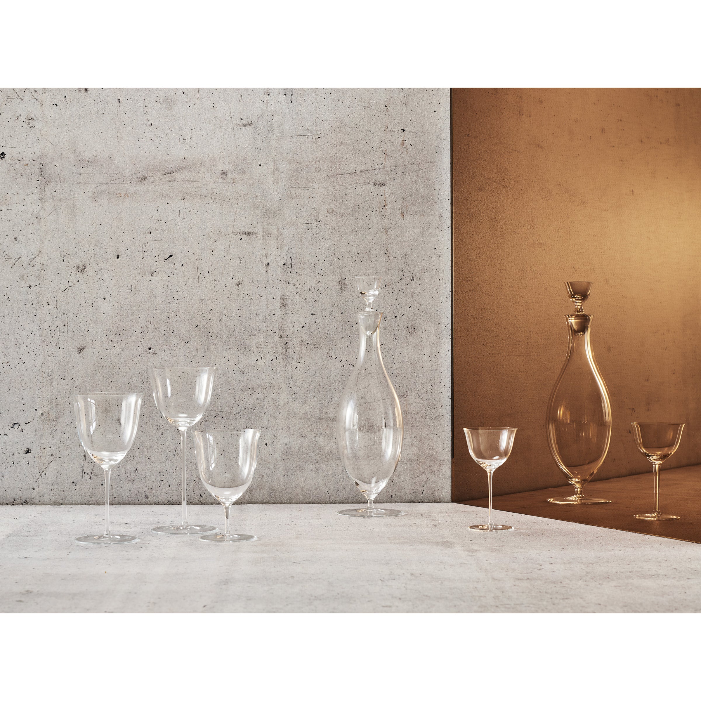 A display of Drinking Set No. 238 - Patrician by Lobmeyr showcases wine glasses, goblets, and elegant crystal decanters. The mouth-blown pieces rest against textured gray and warm brown backgrounds, highlighting their Wiener Werkstätte-inspired artistry.