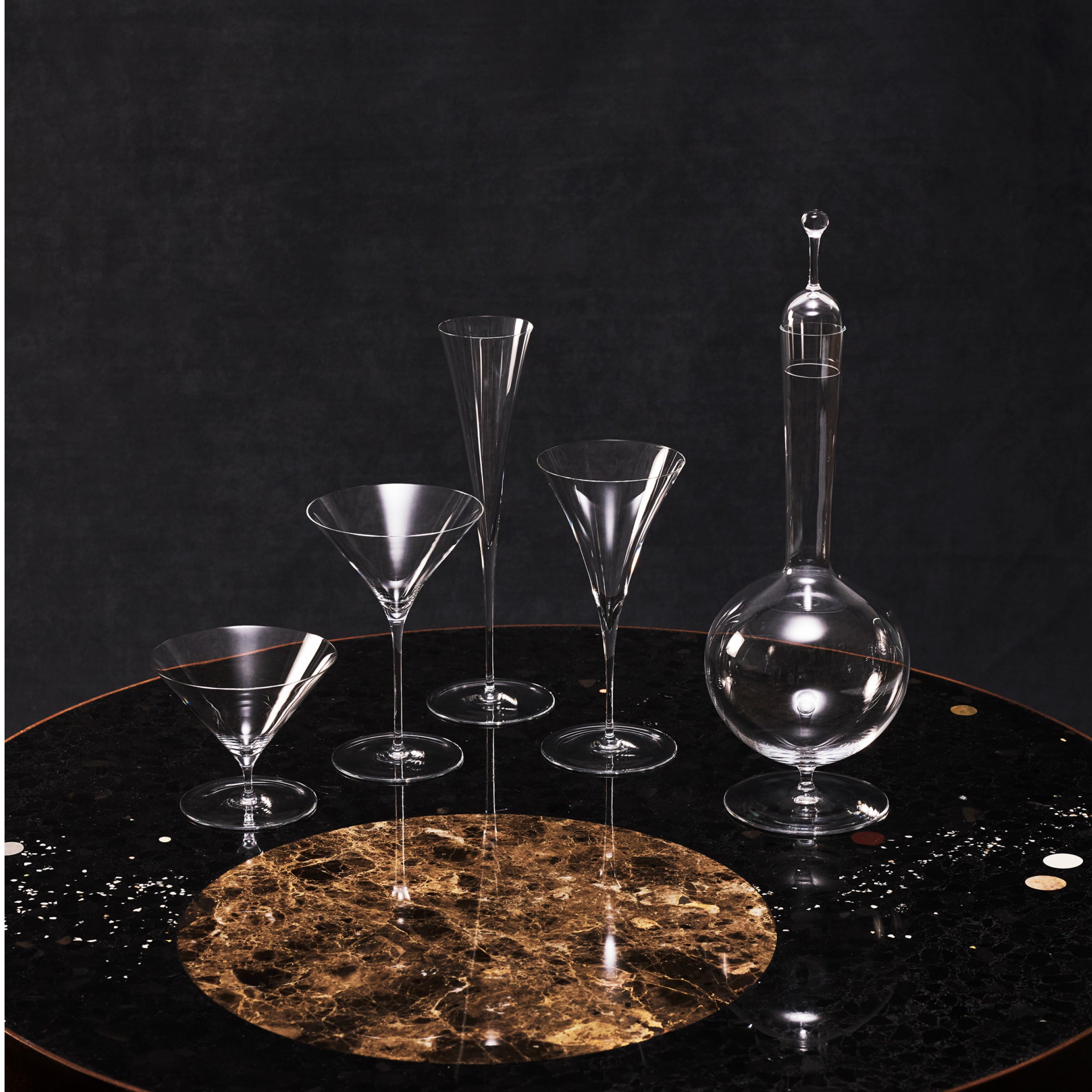 The Lobmeyr Drinking Set No. 240 - Ambassador features five elegant mouth-blown lead-free crystal pieces, including a decanter and various glasses, displayed on a round dark marble table with circular patterns. The solid black backdrop accentuates reflections on the crystal surfaces.