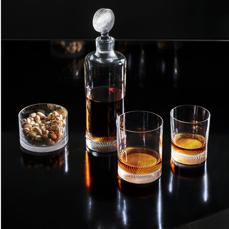 The Lobmeyr Drinking Set No. 248 - Loos, featuring a crystal decanter with an intricate pattern and amber liquid, is paired with two matching glasses on a shiny black surface. A bowl of mixed nuts completes this sophisticated setting inspired by modern glass design by Adolf Loos.