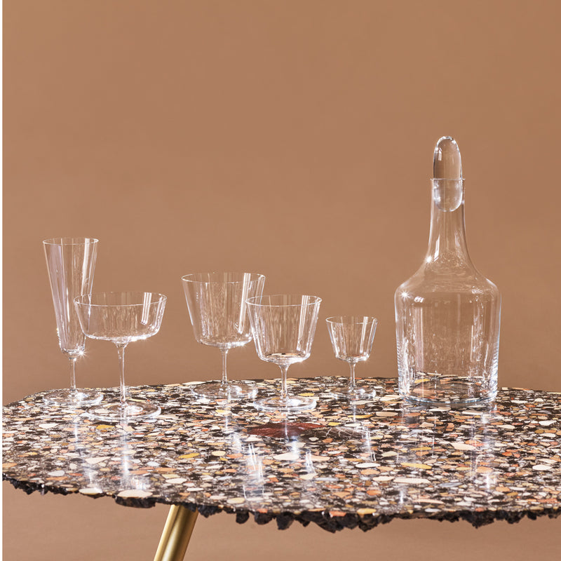 Lobmeyrs Drinking Set No. 257 - Commodore, a collection of mouth-blown lead-free crystal glasses and a decanter designed by Oswald Haerdtl, is elegantly showcased on a polished speckled stone table with a brown backdrop. This series includes wine, champagne, and cocktail glasses.