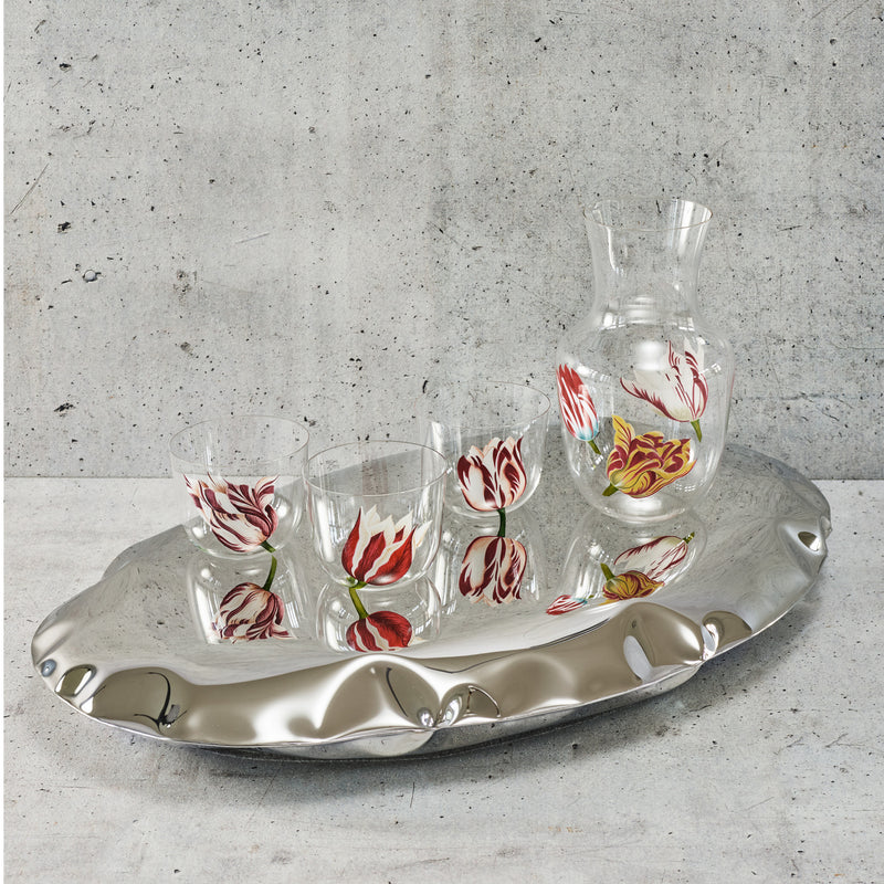 A shiny silver tray displays a Lobmeyr Alpha water pitcher and three Tulipmania tumblers, all featuring red and white tulip blossom designs against a textured gray background. Each lead-free crystal piece ensures elegance with every pour.