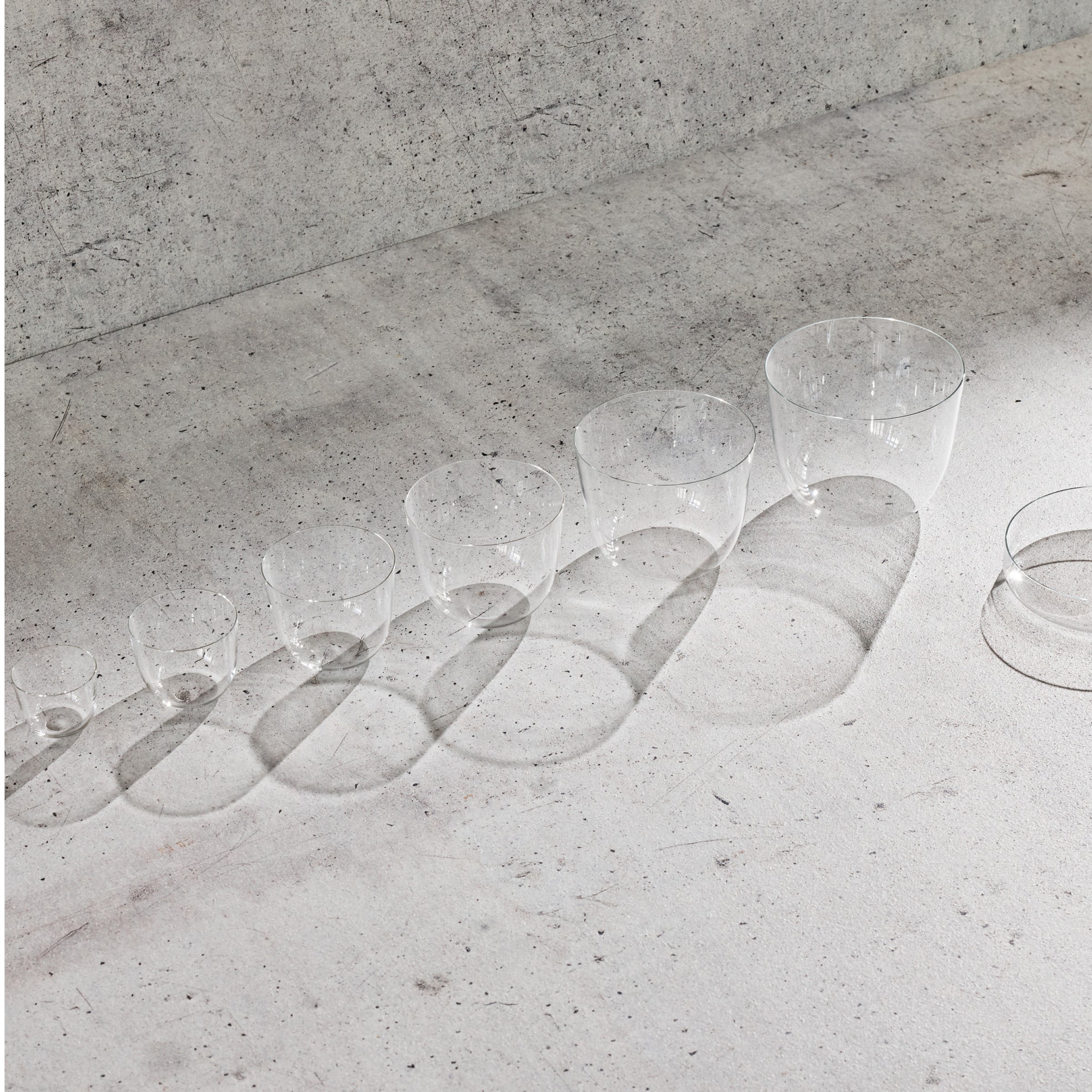 Alpha Cocktail Tumblers by Lobmeyr, crafted from mouth-blown lead-free crystal, sit elegantly on a textured concrete surface. Their transparent cylindrical forms cast elongated shadows, creating a minimalist and artistic visual effect.