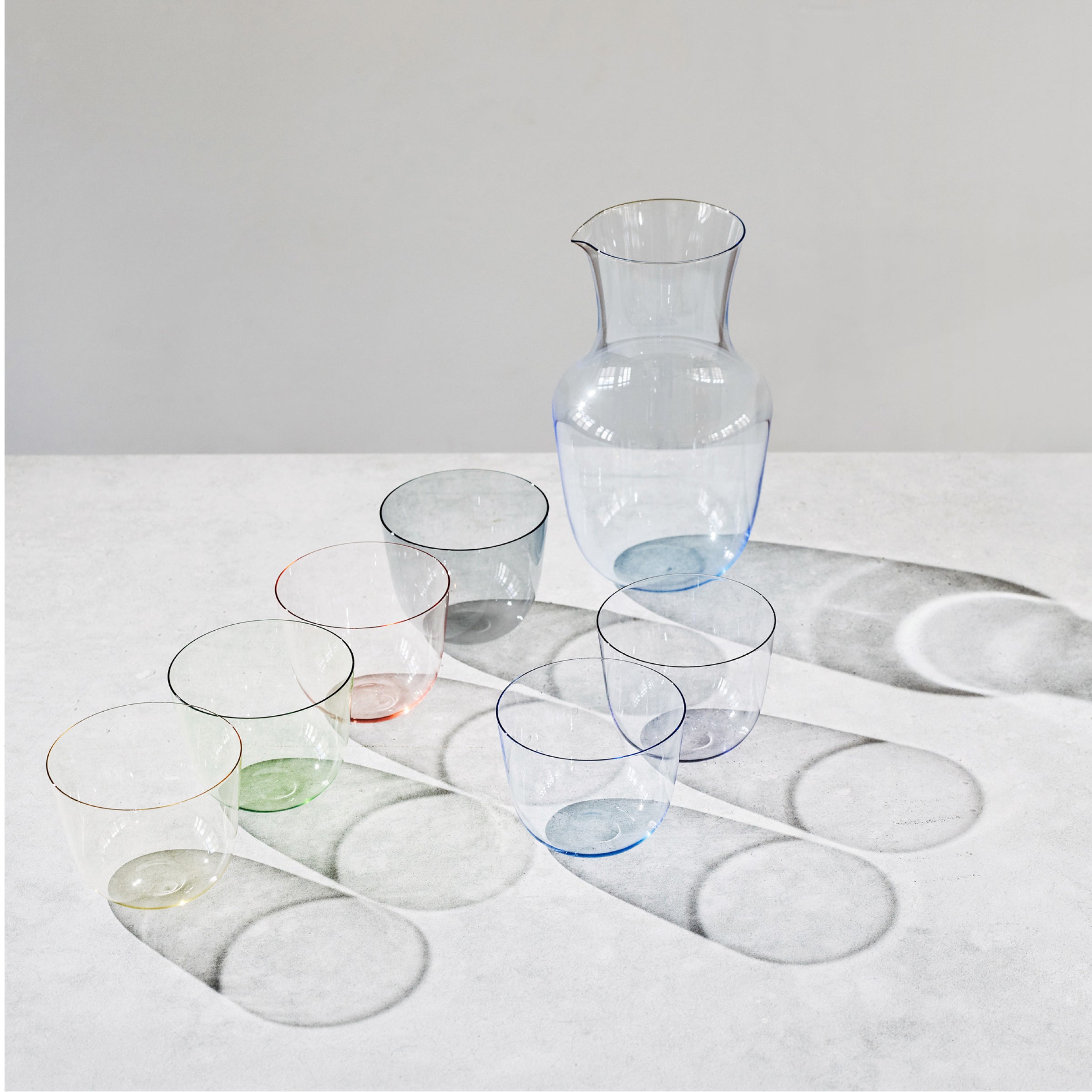 A clear glass Alpha Water Pitcher by Lobmeyr and six stackable drinking glasses with subtle colored rims sit on a white surface, casting soft shadows. The mouth blown crystal glasses, reminiscent of the Alpha Beaker design, are arranged in an irregular pattern that beautifully reflects light.