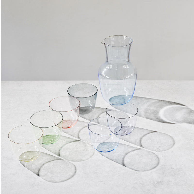 A set of six stackable Alpha Water Tumblers in pastel hues and a matching pitcher, crafted from Lobmeyrs mouth-blown lead-free crystal, are arranged on a light surface. The glasses and Alpha Beaker pitcher create distinct shadow patterns.
