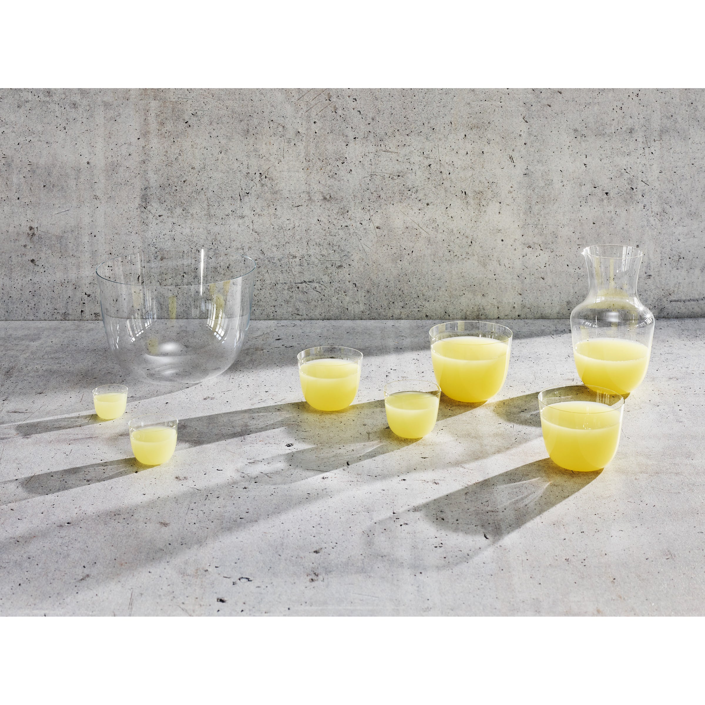 Transparent, stackable cups alongside a Lobmeyr Alpha Water Pitcher filled with yellow liquid rest on a textured concrete surface, casting elongated shadows in the sunlight.