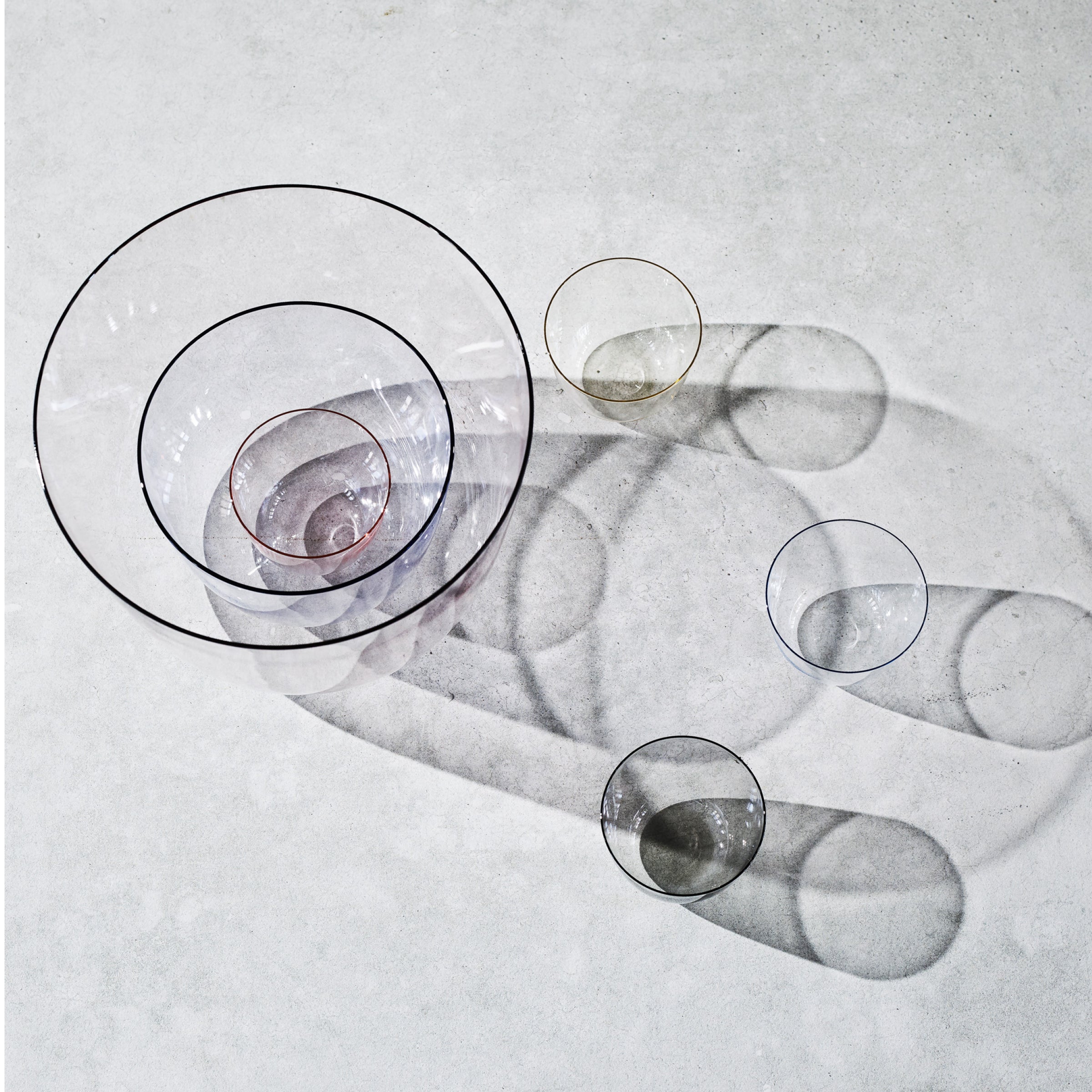 Overhead view of six Alpha Water Tumblers by Lobmeyr, crafted from mouth-blown lead-free crystal, casting shadows on a light gray surface. Arranged in descending size from top left to bottom right, these stackable pieces create intricate overlapping patterns with their delicate form.