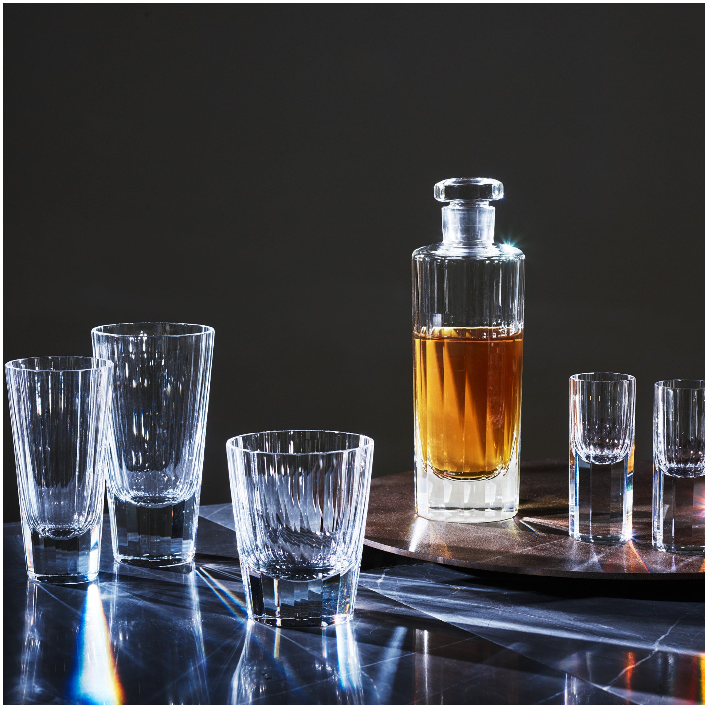 A glass decanter filled with amber liquid sits on a tray surrounded by Lobmeyrs Drinking Set No. 272 - Triennale liqueur glasses, their lead-free crystal surfaces reflecting light against a dark background.