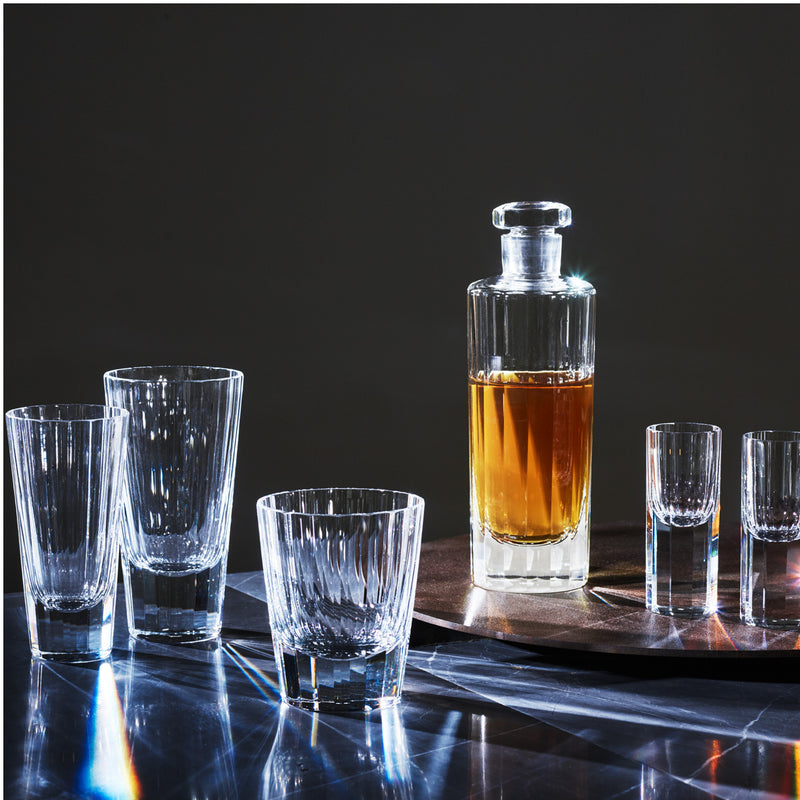 A glass decanter filled with amber liquid sits on a tray surrounded by Lobmeyrs Drinking Set No. 272 - Triennale liqueur glasses, their lead-free crystal surfaces reflecting light against a dark background.