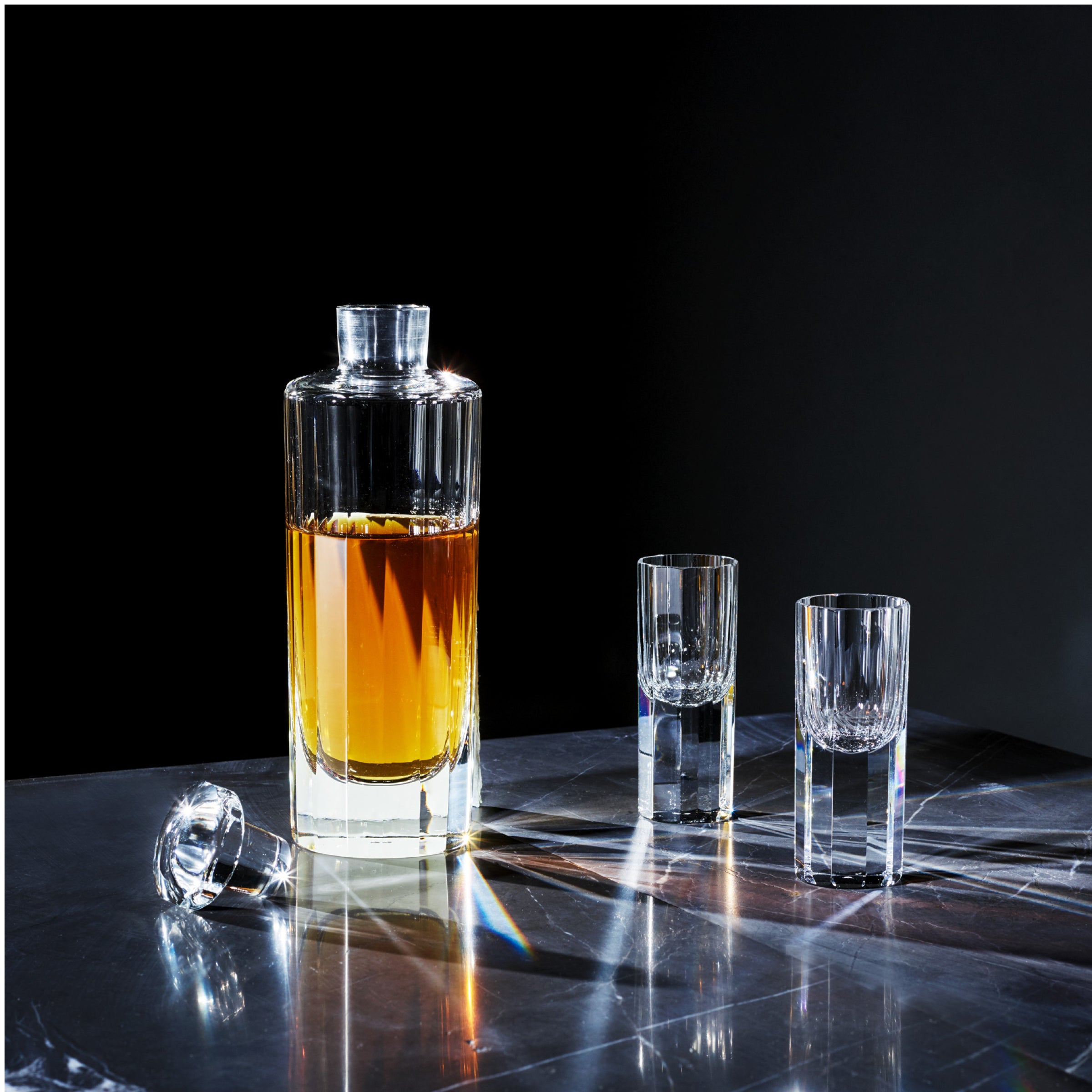 A glass decanter filled with amber liquid is on a reflective surface, surrounded by two empty Lobmeyr Drinking Set No. 272 - Triennale liqueur glasses. The decanters stopper rests on the table, while light dances with reflections and shadows across the elegant crystal barware.