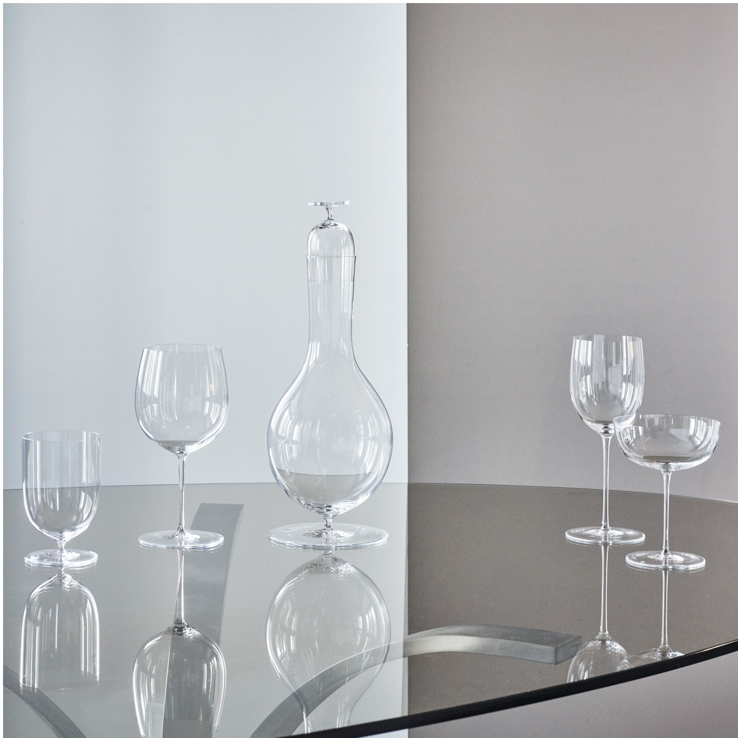 A set of Lobmeyrs Drinking Set No. 280 - Wiener Gemischter Satz is displayed on a round glass table, including a tall decanter, two crystal wine glasses, one shorter glass, and two cocktail glasses. The softly lit background is an elegant blend of white and gray tones.