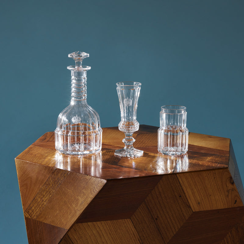 A clear glass decanter, a tall decorative-stemmed glass, and a short tumbler rest on a geometric wooden table against a blue backdrop. The intricate etched designs of the crystal glasses from Lobmeyrs Drinking Set No. 2 - Hofburg reflect light beautifully, highlighting exceptional craftsmanship.