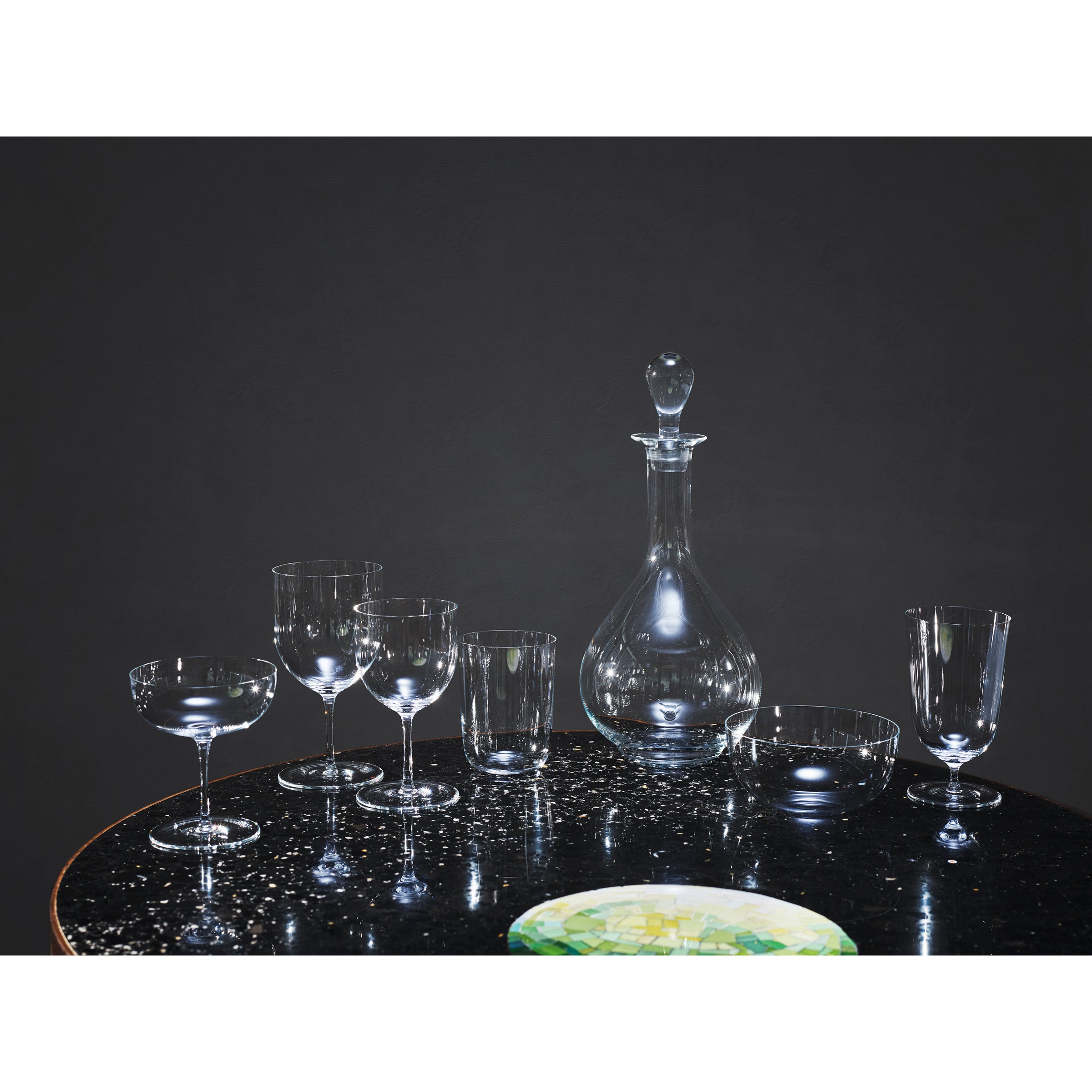 The Lobmeyr Drinking Set No. 4, crafted in crystal glass with a decanter, wine glasses, and tumblers in muslin glass style, is elegantly displayed on a glossy black table. A small multicolored plate stands out against the plain dark gray backdrop.