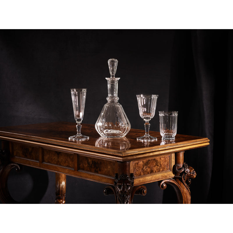 A dark wooden table with ornate legs elegantly showcases a mouth-blown crystal decanter and three pieces from the Lobmeyr Drinking Set No. 98 - Palais with Facette Cut, all set against a black background.