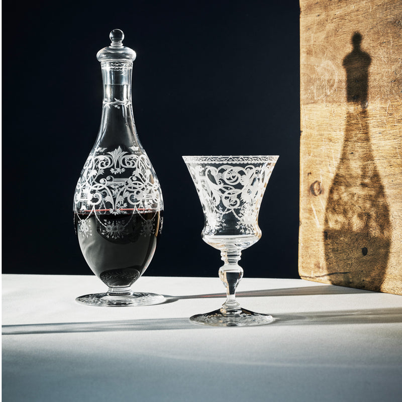 A glass decanter with red wine is next to a Lobmeyr Drinking Set No. 231 - Barock, featuring engraved ornament, on a white surface. The intricate patterns cast shadows on a wooden backdrop, as dramatic lighting highlights their elegance.