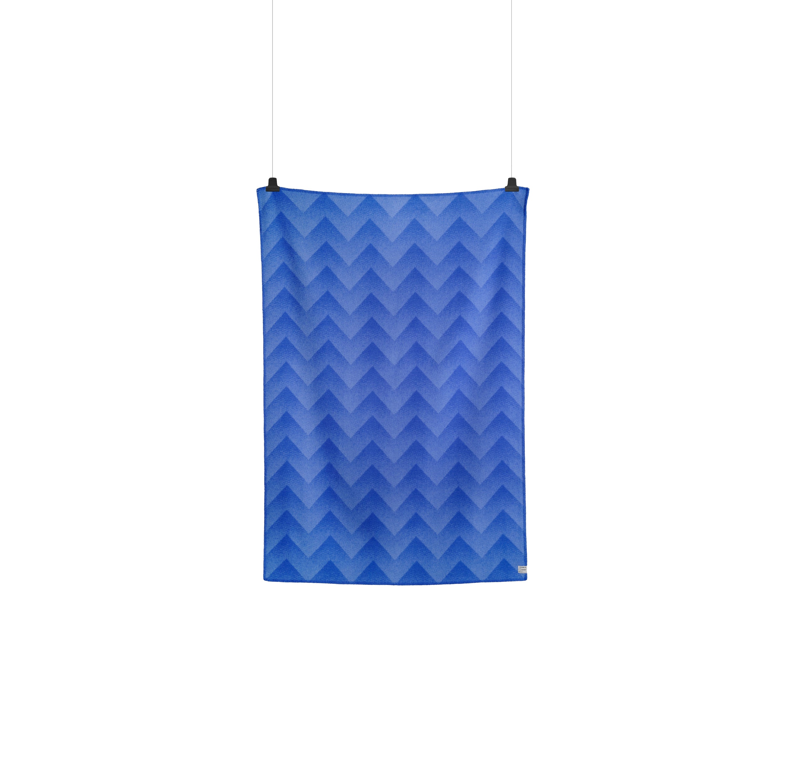The Roros Lynild Throw, with its chevron pattern in blue, reminiscent of Anderssen & Volls fire stitch technique, elegantly hangs against a pristine white backdrop.