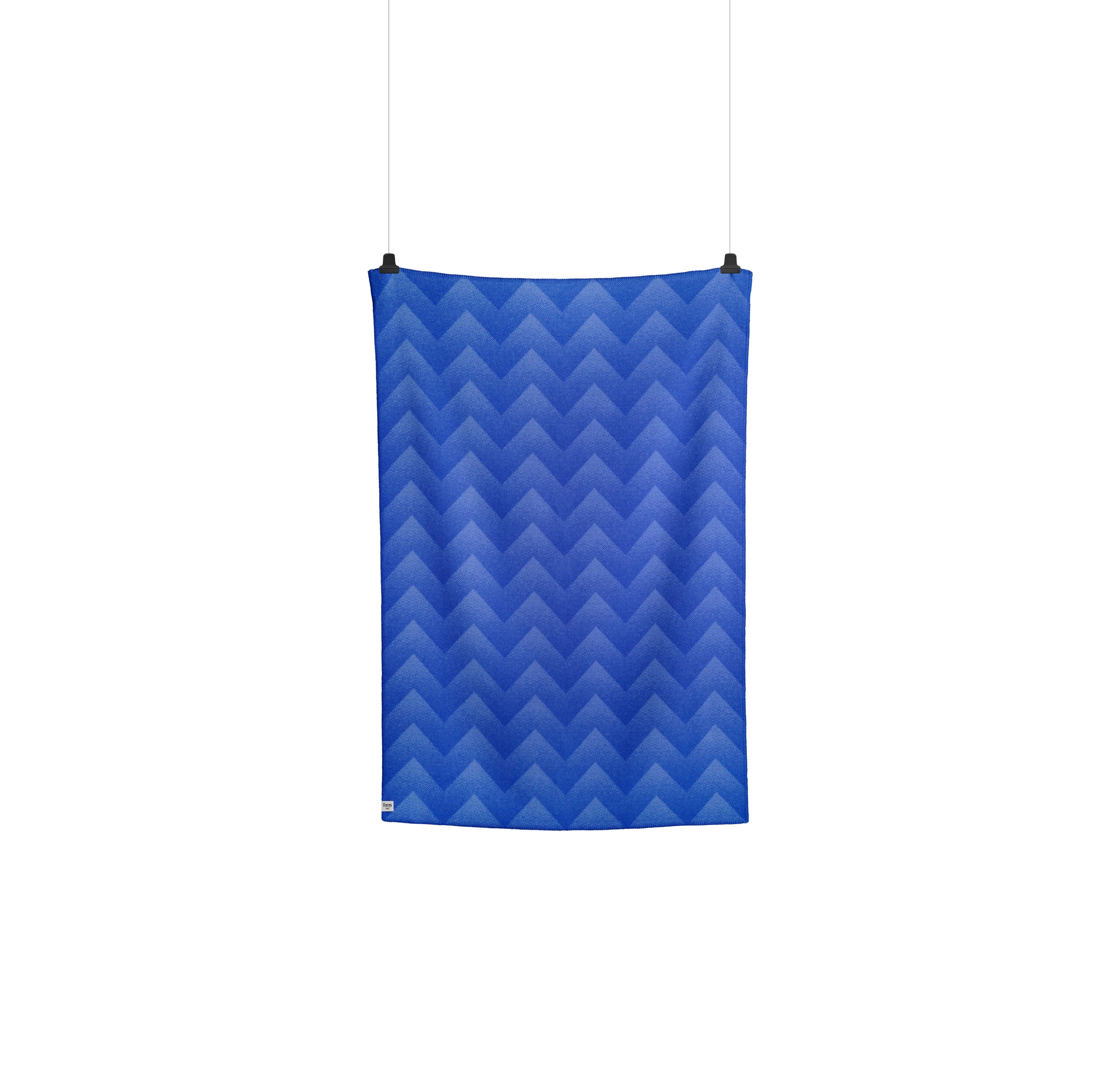 The Lynild Throw by Roros, with its intricate fire stitch-inspired zigzag pattern in blue, hangs vertically against a white background.