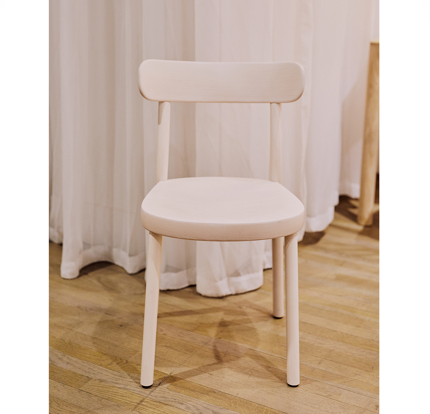 The Chair La Zitta - INVENTORY by TON, a light wooden dining chair with bentwood style and simple design, rests on a polished floor. White curtains in the background contribute to an elegant, airy setting, reflecting classic TON craftsmanship.