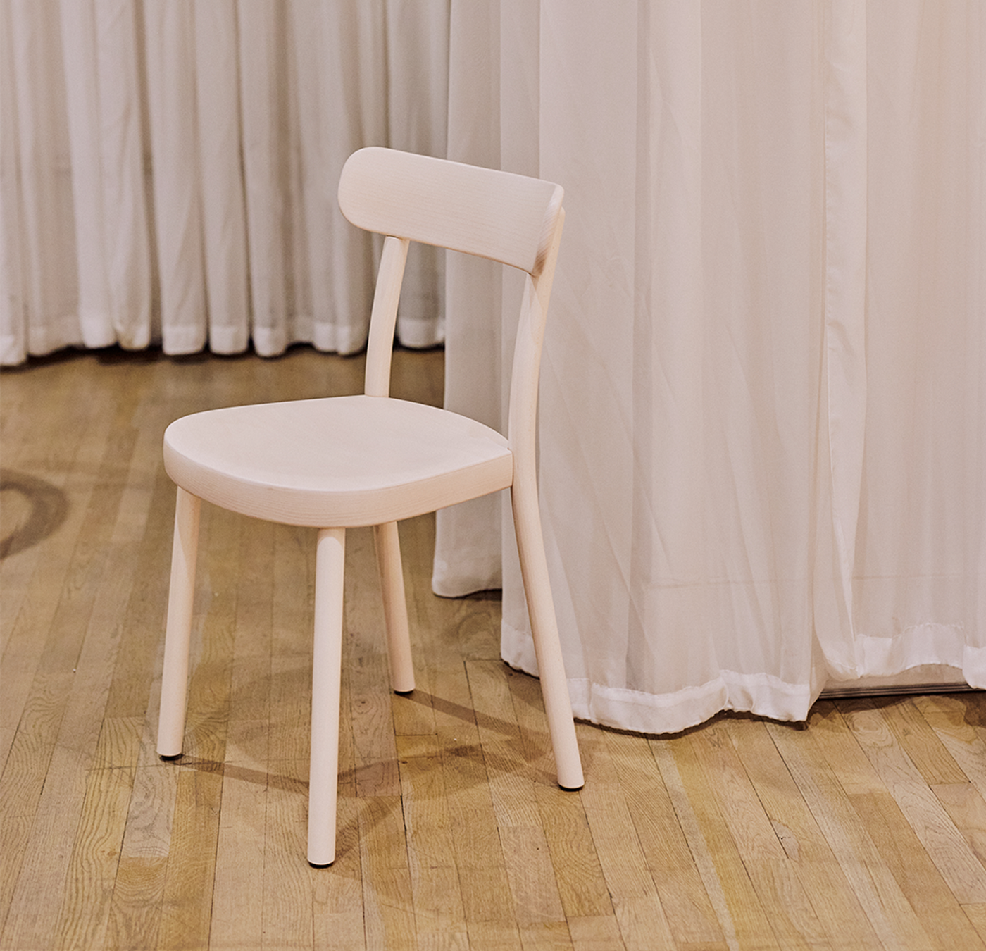 The Chair La Zitta by TON, a simple and light bentwood dining chair with a rounded seat and low backrest, sits on a polished wooden floor. The background features floor-length white curtains, adding an elegant touch reminiscent of a true TON piece.
