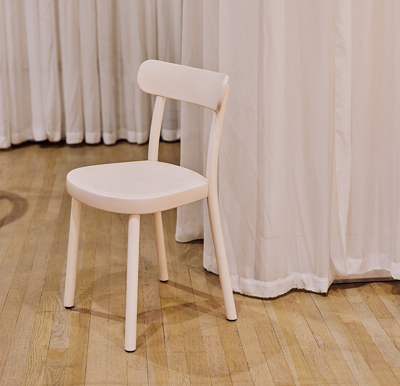 The Chair La Zitta by TON, a simple and light bentwood dining chair with a rounded seat and low backrest, sits on a polished wooden floor. The background features floor-length white curtains, adding an elegant touch reminiscent of a true TON piece.