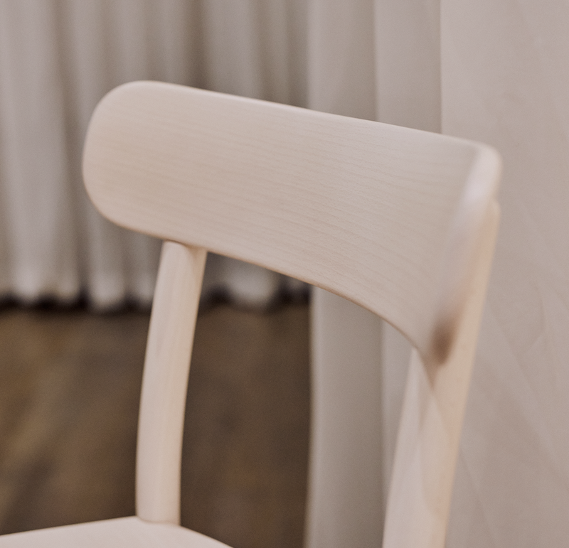 A close-up of Chair La Zitta by TON shows its bentwood design and curved backrest, set against beige curtains and a brown wooden floor, creating a warm ambiance.