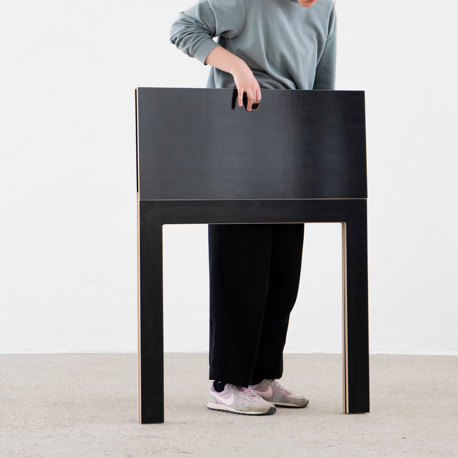 A person stands inside Moormanns Last Minute, a large, hollow, rectangular birch plywood frame, reaching through an opening and creating the illusion of merging with this space-saving furniture. They wear a blue long-sleeve shirt, black pants, and white sneakers.
