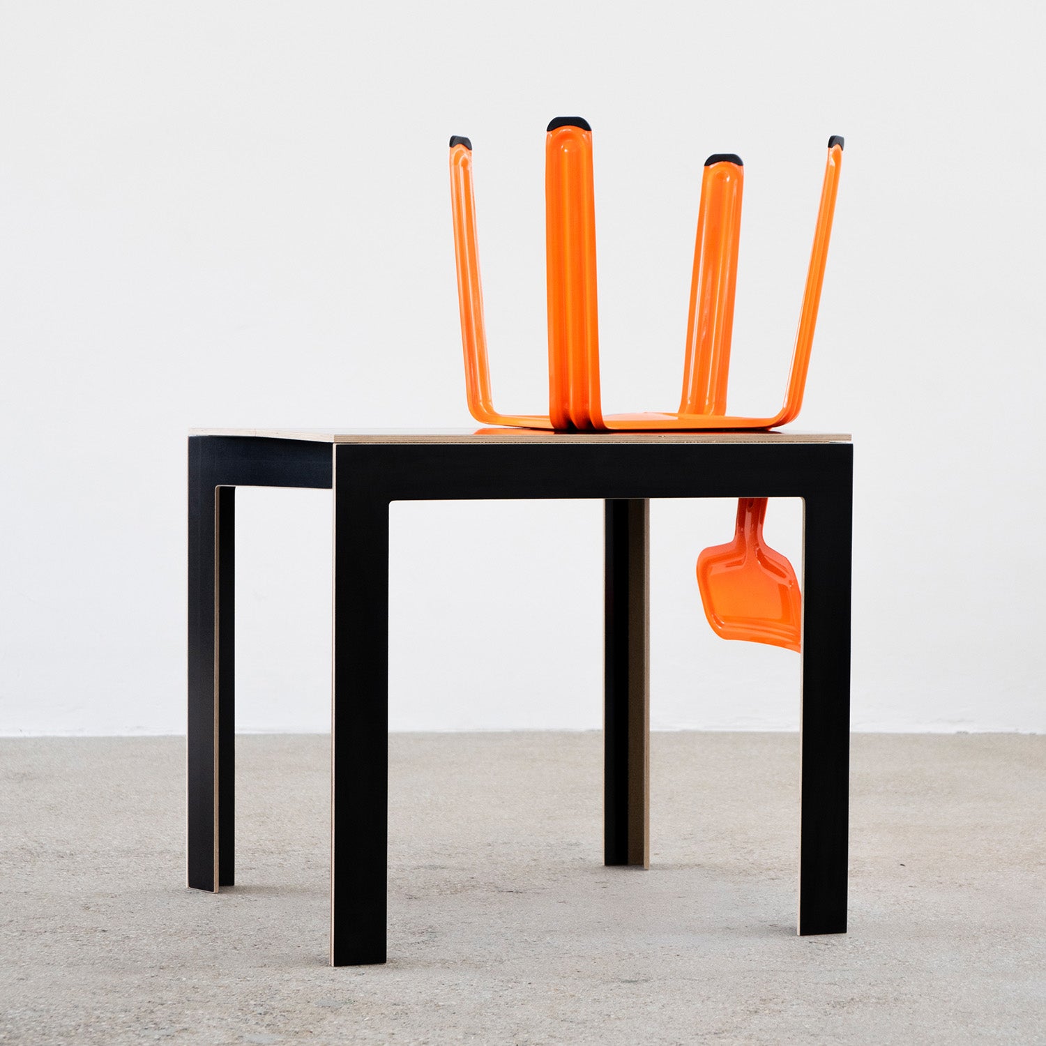 The Moormann Last Minute foldable table, featuring a black rectangular design with wooden edges, stands on a gray floor. An orange chair creatively positioned upside down atop it resembles an abstract sculpture, with its legs extending upward and outward.