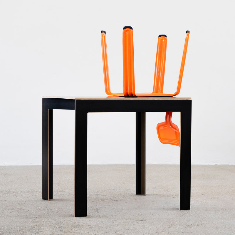 The Moormann Last Minute foldable table, featuring a black rectangular design with wooden edges, stands on a gray floor. An orange chair creatively positioned upside down atop it resembles an abstract sculpture, with its legs extending upward and outward.