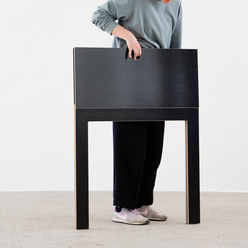 A person stands inside Moormanns Last Minute, a large, hollow, rectangular birch plywood frame, reaching through an opening and creating the illusion of merging with this space-saving furniture. They wear a blue long-sleeve shirt, black pants, and white sneakers.