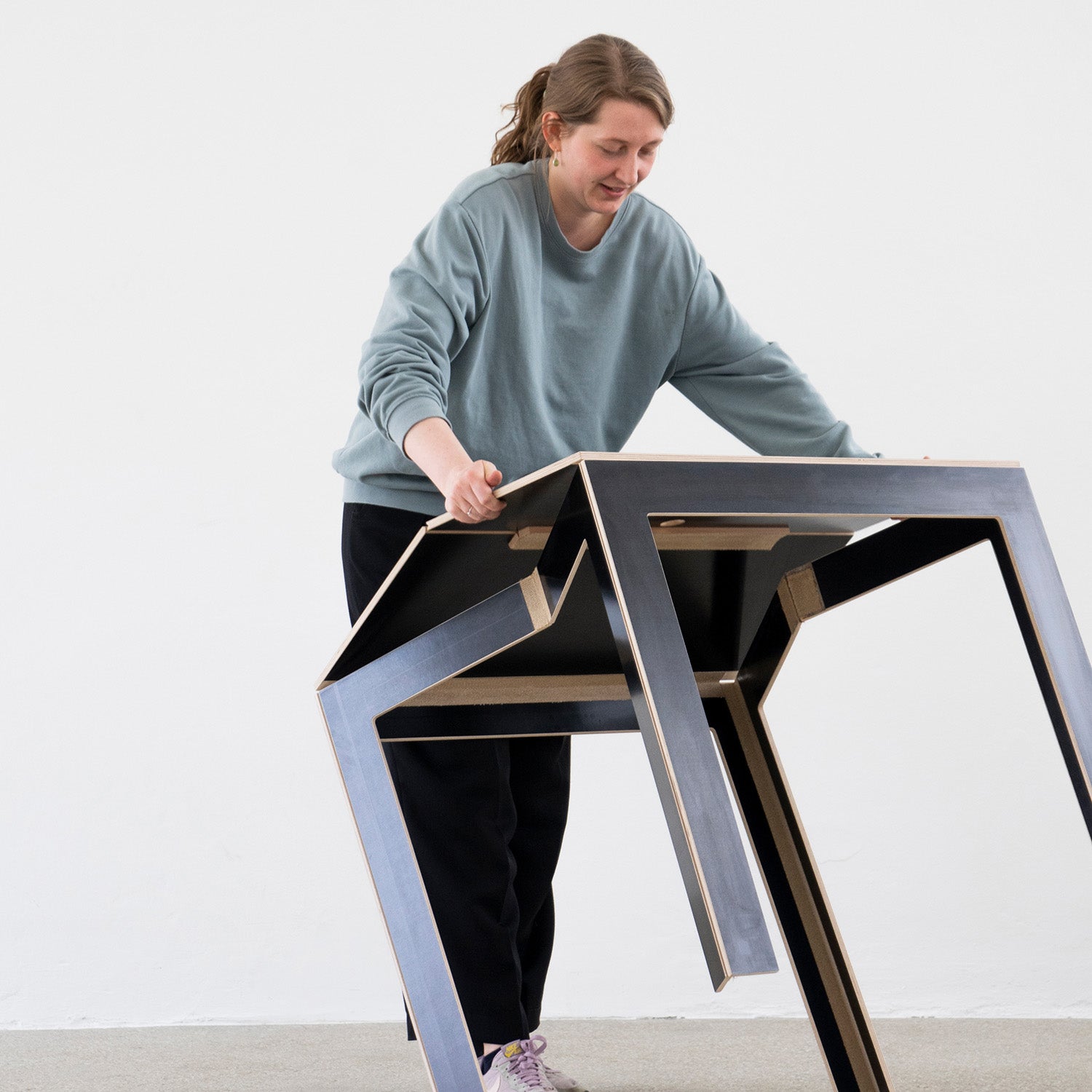 Wearing a blue sweater and black pants, a person skillfully folds the Last Minute table by Moormann. With its geometric birch plywood design and zigzag legs, this table blends perfectly against a white wall, providing an exceptional space-saving solution.