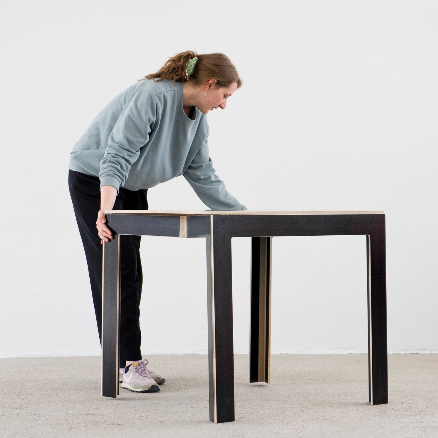 Wearing a light blue sweater, black pants, and sneakers, a person with a ponytail assembles the Moormann Last Minute black foldable table with wooden edges. The space-saving furniture fits perfectly against the minimalistic white background.