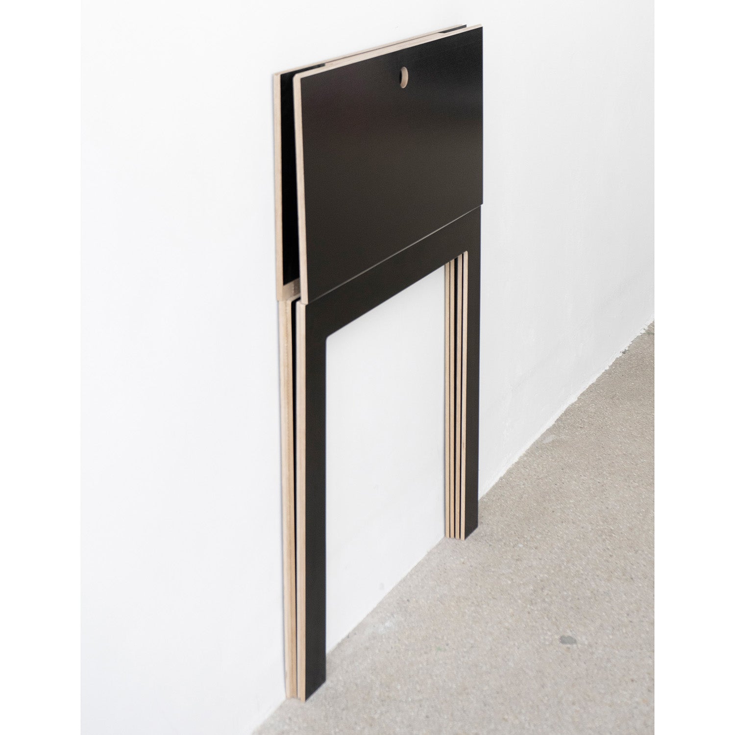 A black birch plywood folding table, the Last Minute by Moormann, rests flat against a white wall, showcasing its minimalist design and wooden edges. This sleek space-saving piece features a small circular cutout near the top, beautifully complementing the neutral gray floor.